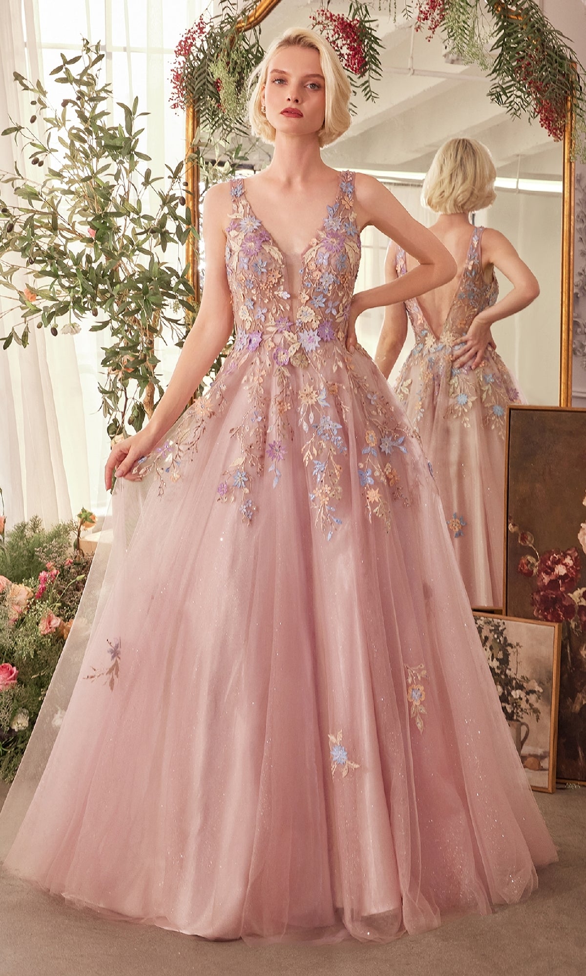 A woman in the Mauve Pink Long Prom Ball Gown (CLA1394), featuring floral-lace appliques, stands before a mirror amid lush greenery and blooming flowers.