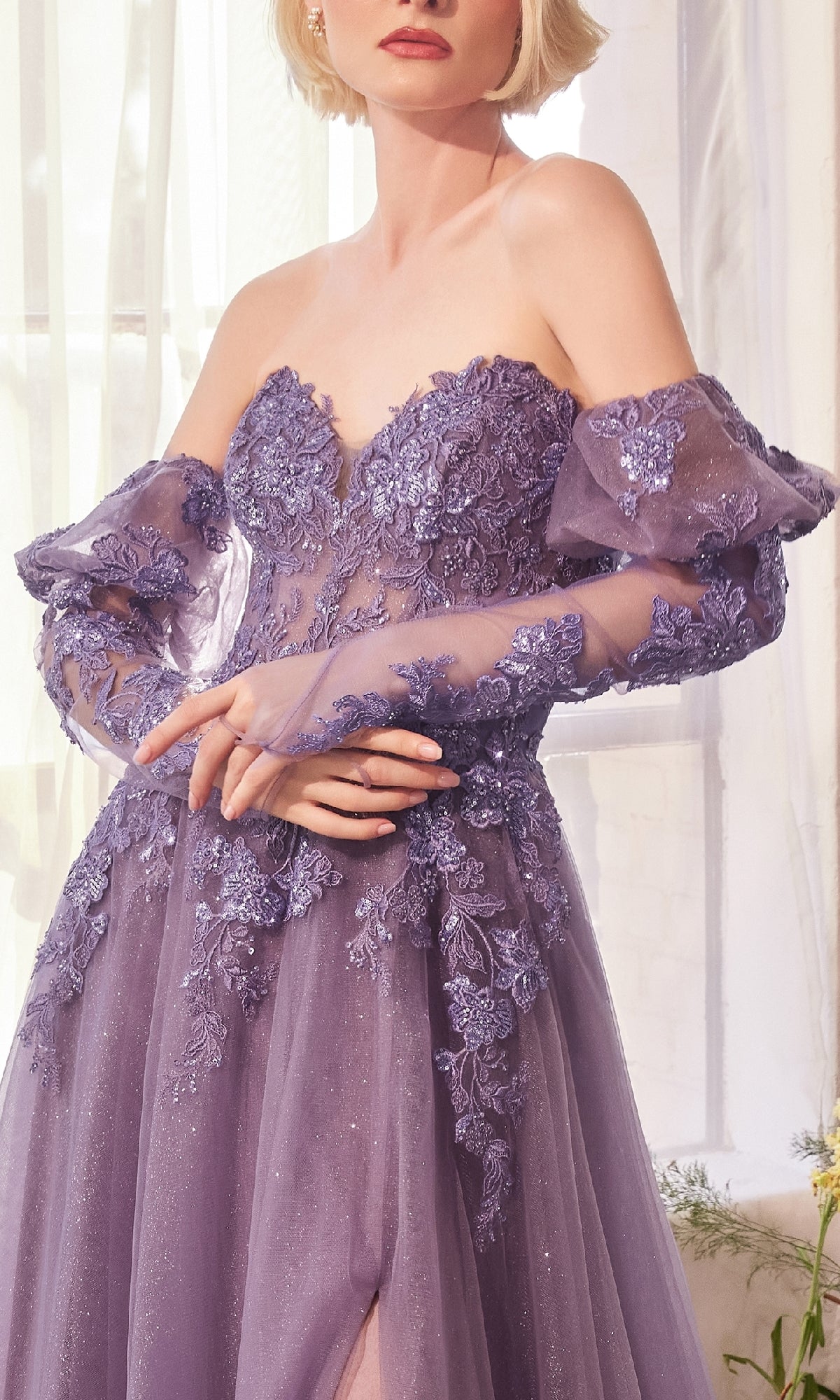 A woman stands indoors near a window wearing the Andrea & Leo A1398 Strapless Glitter Prom Gown.
