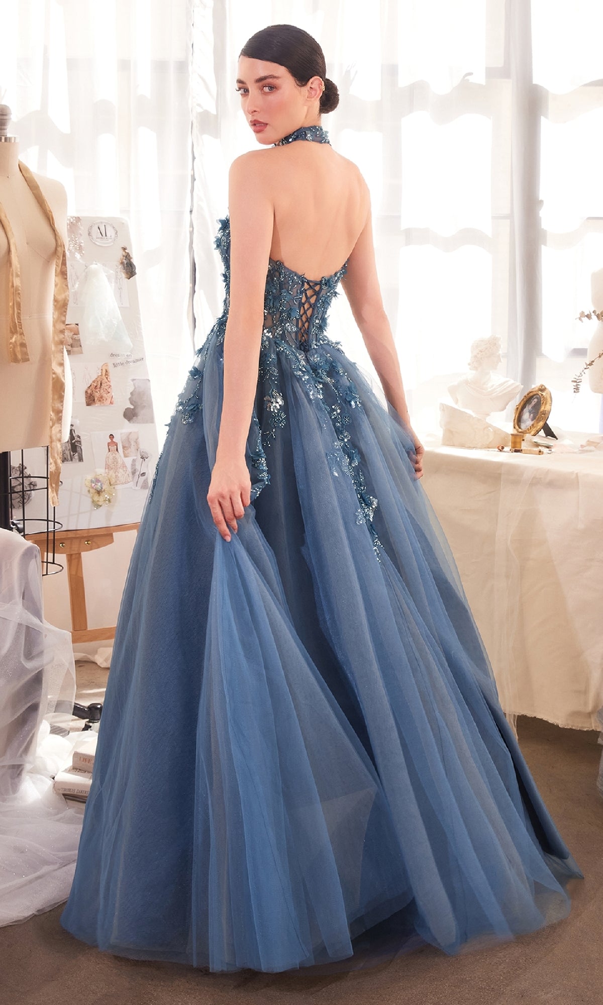 A woman stands in a sunlit room wearing the Strapless Floral Prom Gown: Andrea & Leo A1399, a blue, floor-length dress with floral appliqué and lace-up back details.
