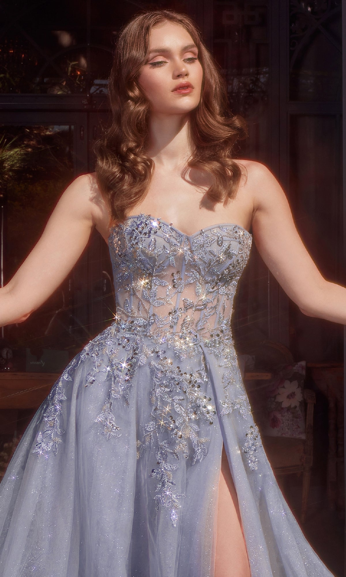 A woman in the Andrea & Leo A1415 Strapless Prom Ball Gown—a light blue floral-embellished evening dress with a sheer bodice and thigh-high slit—stands gracefully with her arms slightly outstretched.
