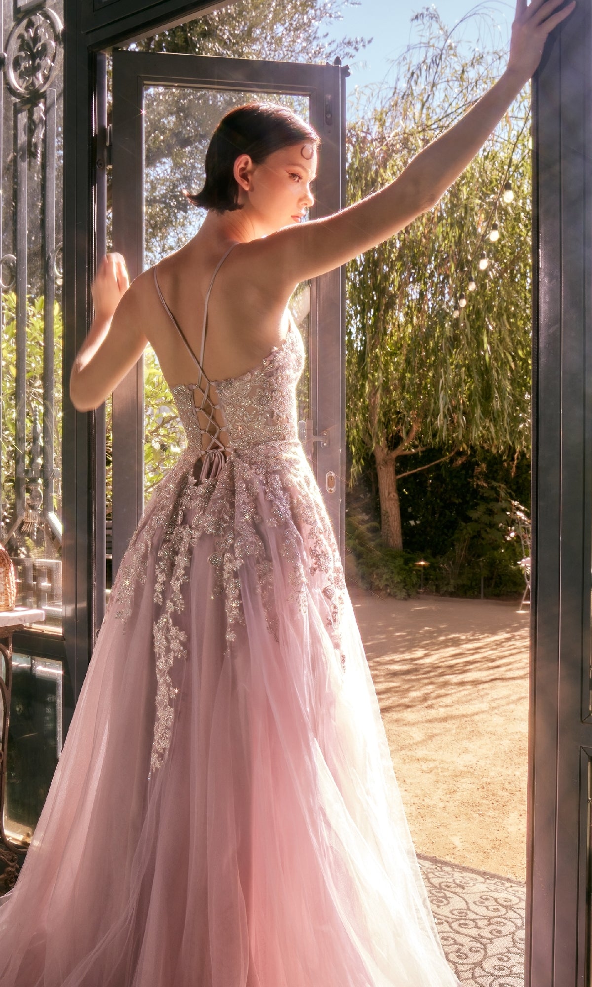 A woman wearing the Andrea & Leo A1416 Lace-Up Long A-Line Prom Dress in pink, adorned with beads, stands in an open doorway and gazes toward a sunlit garden.