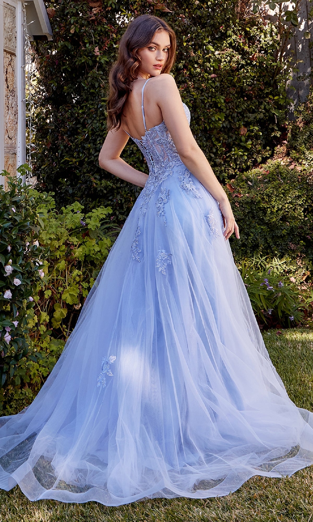 Outdoors, a woman wears the Embroidered Long Prom Dress: Andrea & Leo A1417—a light blue, floor-length gown with an embroidered bodice, floral embellishments, and thin straps—as she looks over her shoulder.