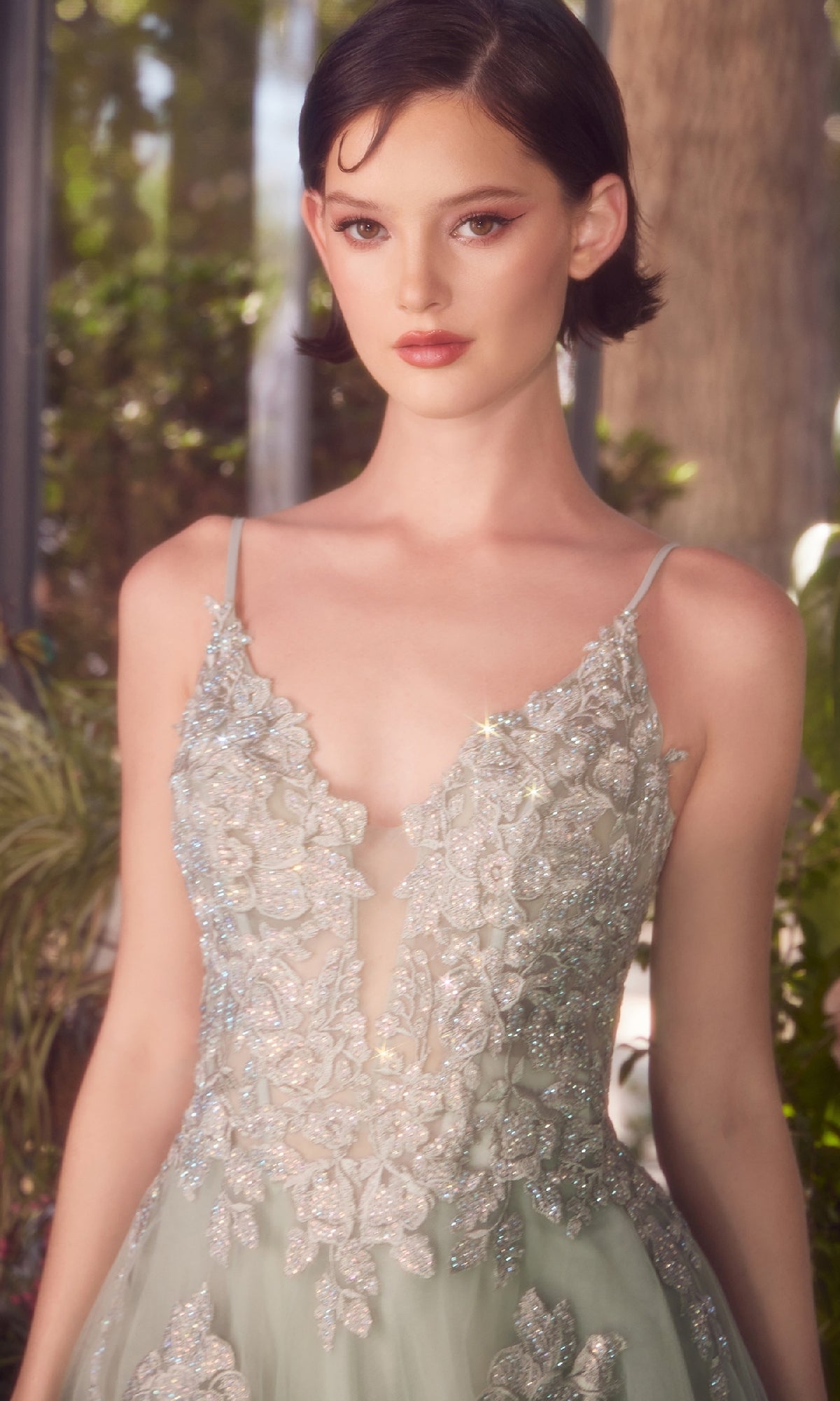 A woman wearing the Andrea & Leo A1417 embroidered long prom dress with an a-line silhouette and thin straps stands in a softly lit indoor garden.