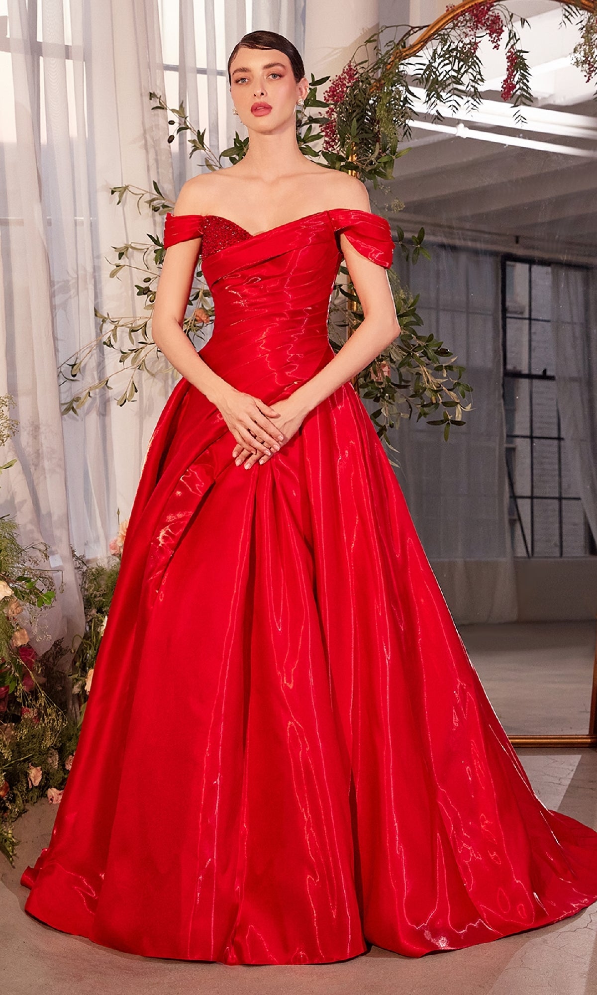 A woman stands indoors wearing the Off-the-Shoulder Prom Ball Gown: Andrea & Leo A1424, a floor-length bright red formal dress with a fitted bodice and full, sweeping skirt.