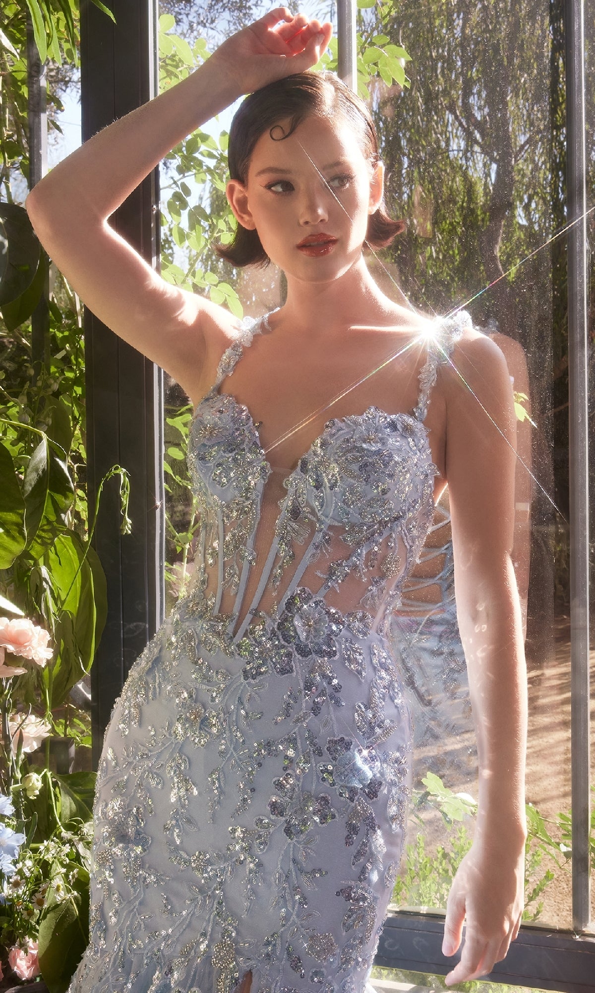A woman stands by a window in natural light wearing the Andrea & Leo A1429 long blue sequin prom dress, featuring sheer panels and floral embellishments.