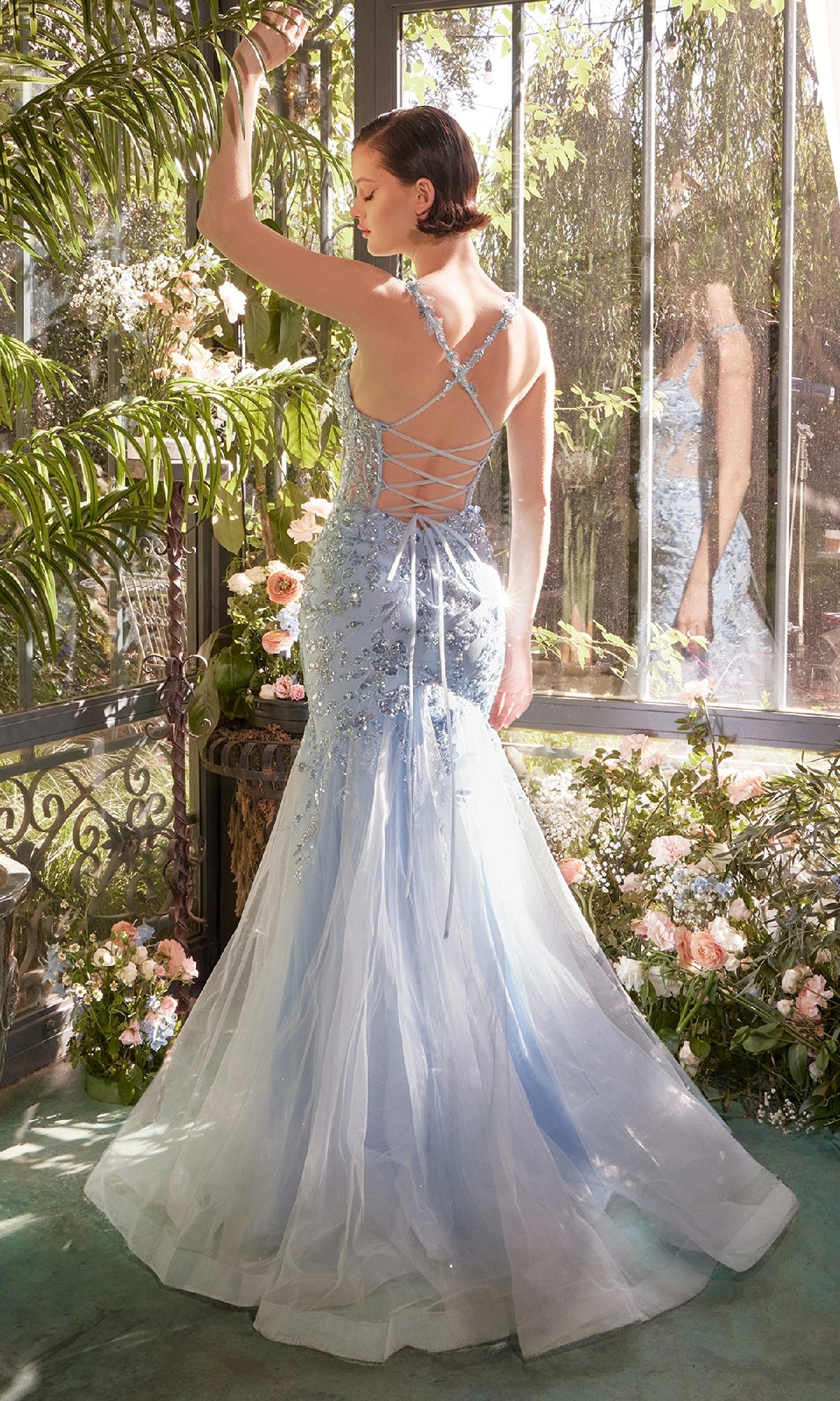 A woman stands among flowers in a sunlit glass conservatory, wearing the Andrea & Leo A1429 Long Blue Sequin Prom Dress—a strappy-back mermaid evening gown that flows gracefully as she is seen from behind.