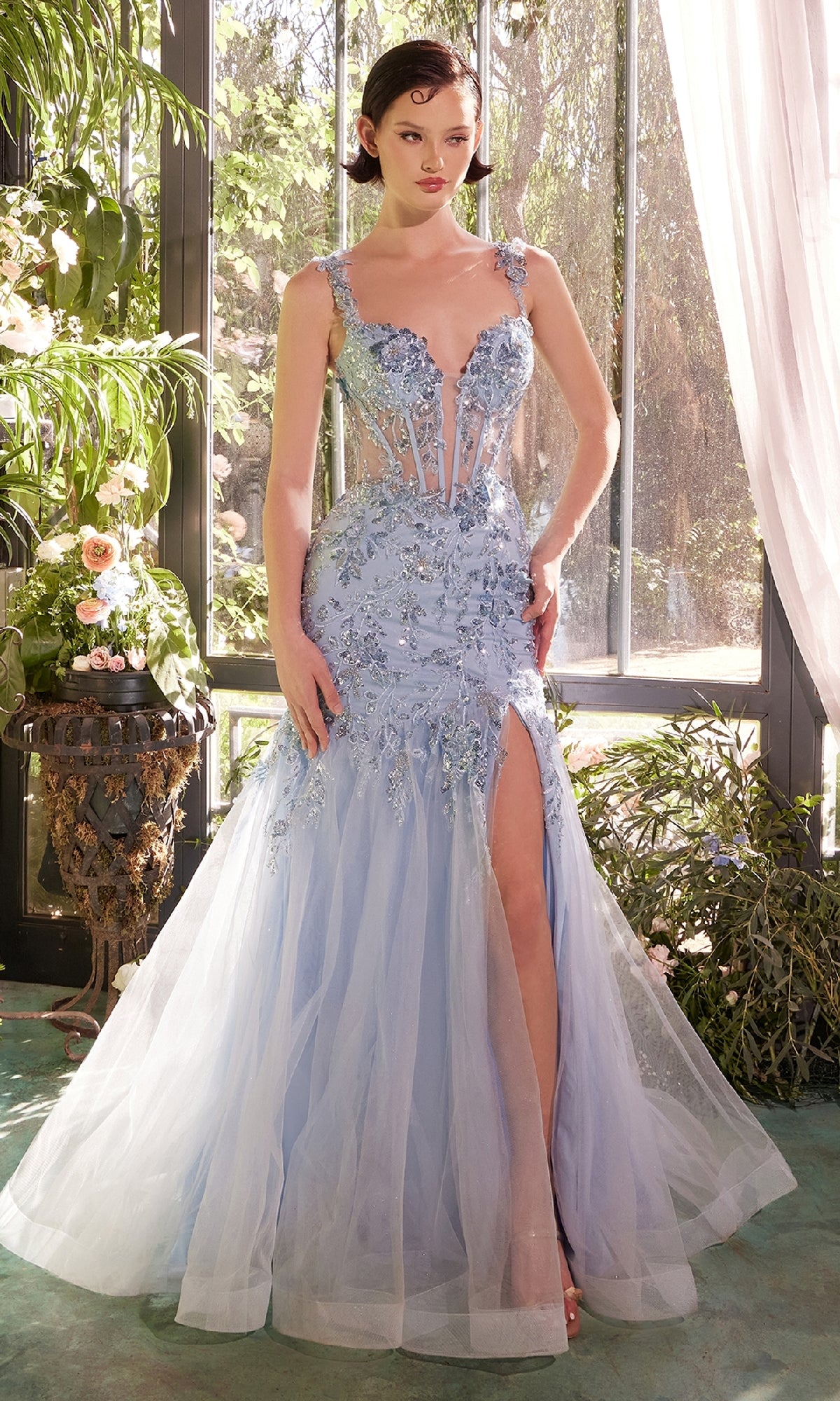 A woman stands indoors near large windows and lush green plants, wearing the Andrea & Leo A1429 Long Blue Sequin Prom Dress with floral embellishments and a thigh-high slit.