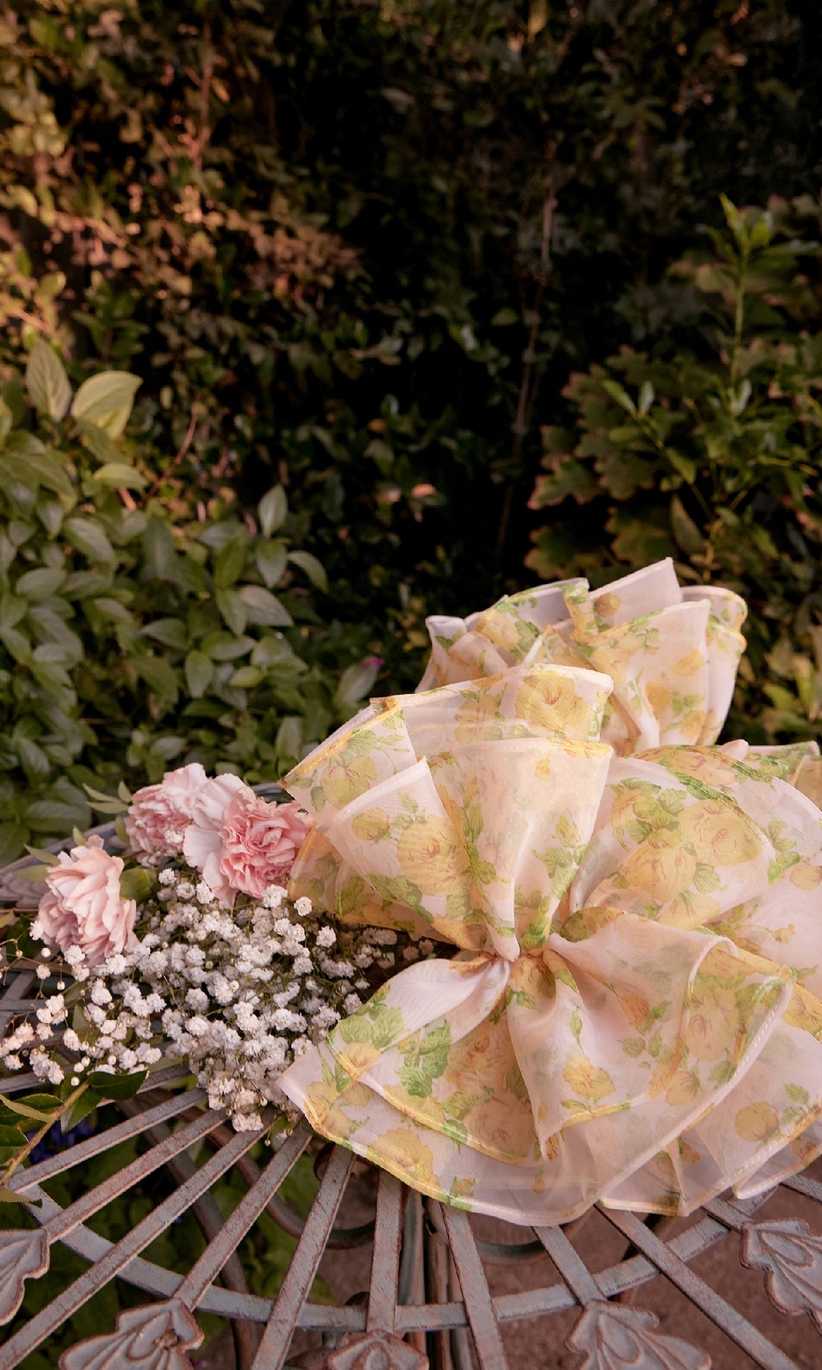 A large, sheer floral bow and a bouquet of pink and white flowers rest on a metal outdoor table, complementing the Andrea & Leo A1430 Yellow Flora-Print Prom Gown against a backdrop of green foliage.