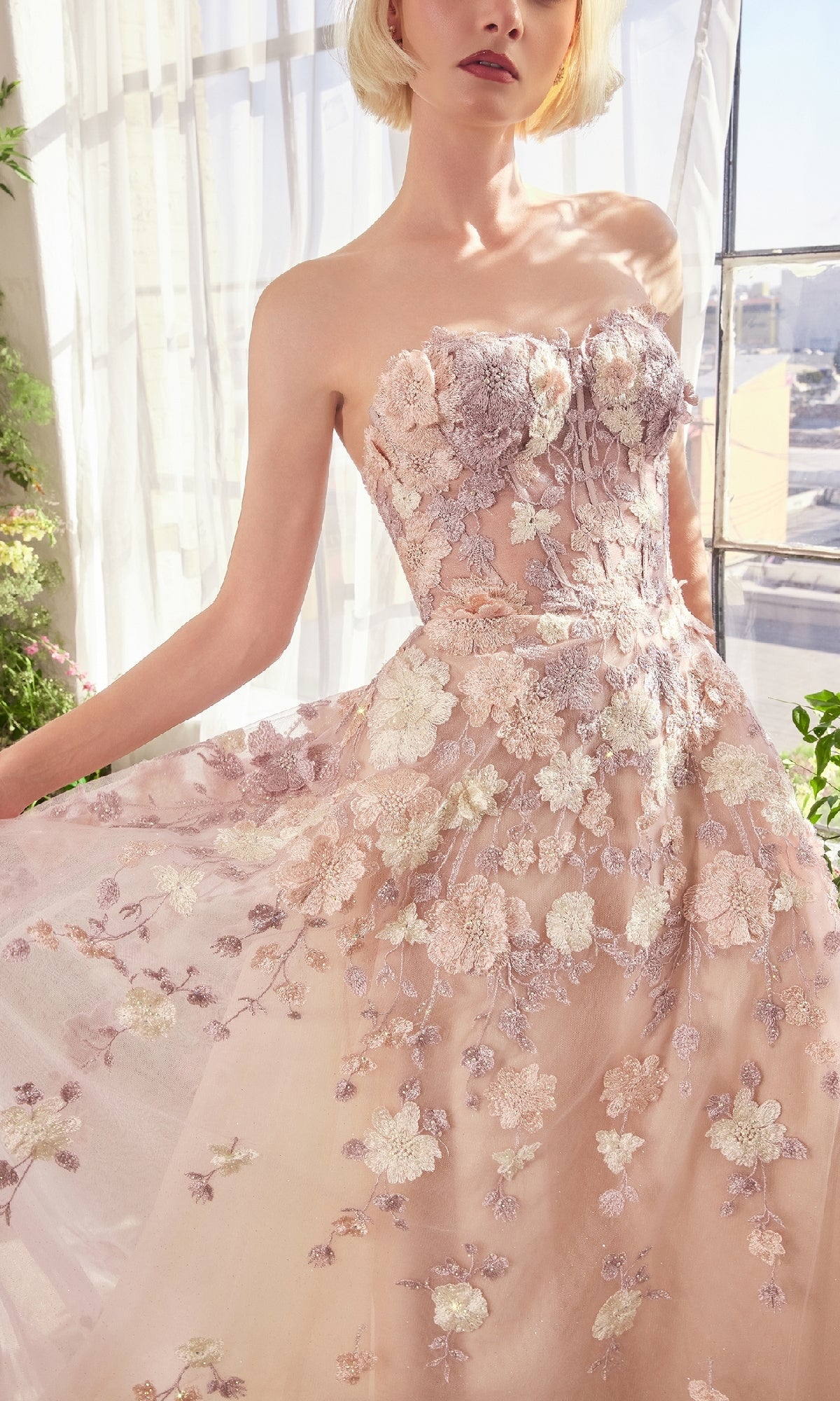 A woman in the Strapless Long Mauve Prom Gown: Andrea & Leo A1453 with 3D floral embroidery stands by a sunlit window, highlighting the elegance of this stunning evening dress.