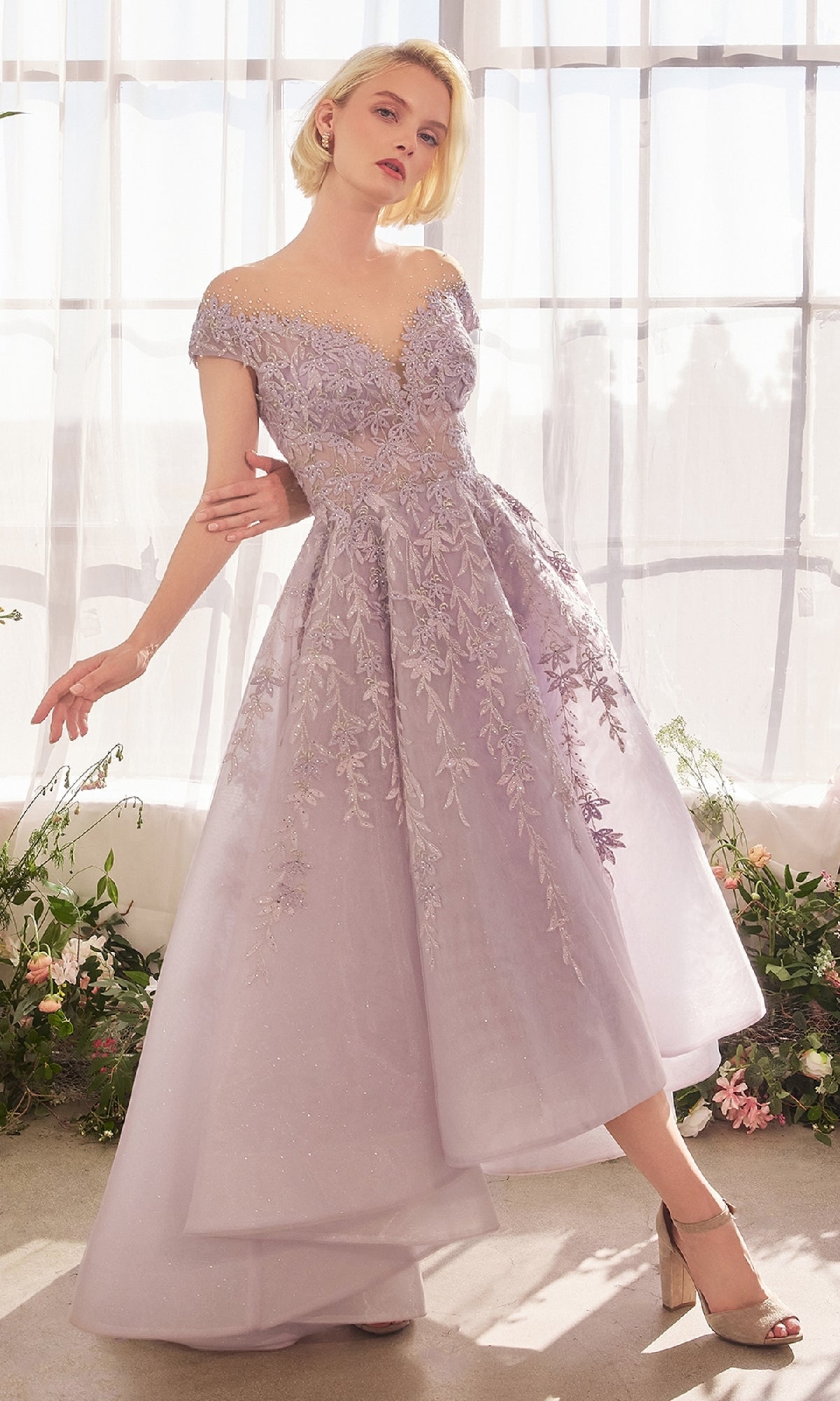 A woman poses with one arm extended in front of sheer curtains and flowers, wearing the Andrea and Leo A1454 long prom dress featuring floral embroidery, a pastel hue, and a high-low hemline.