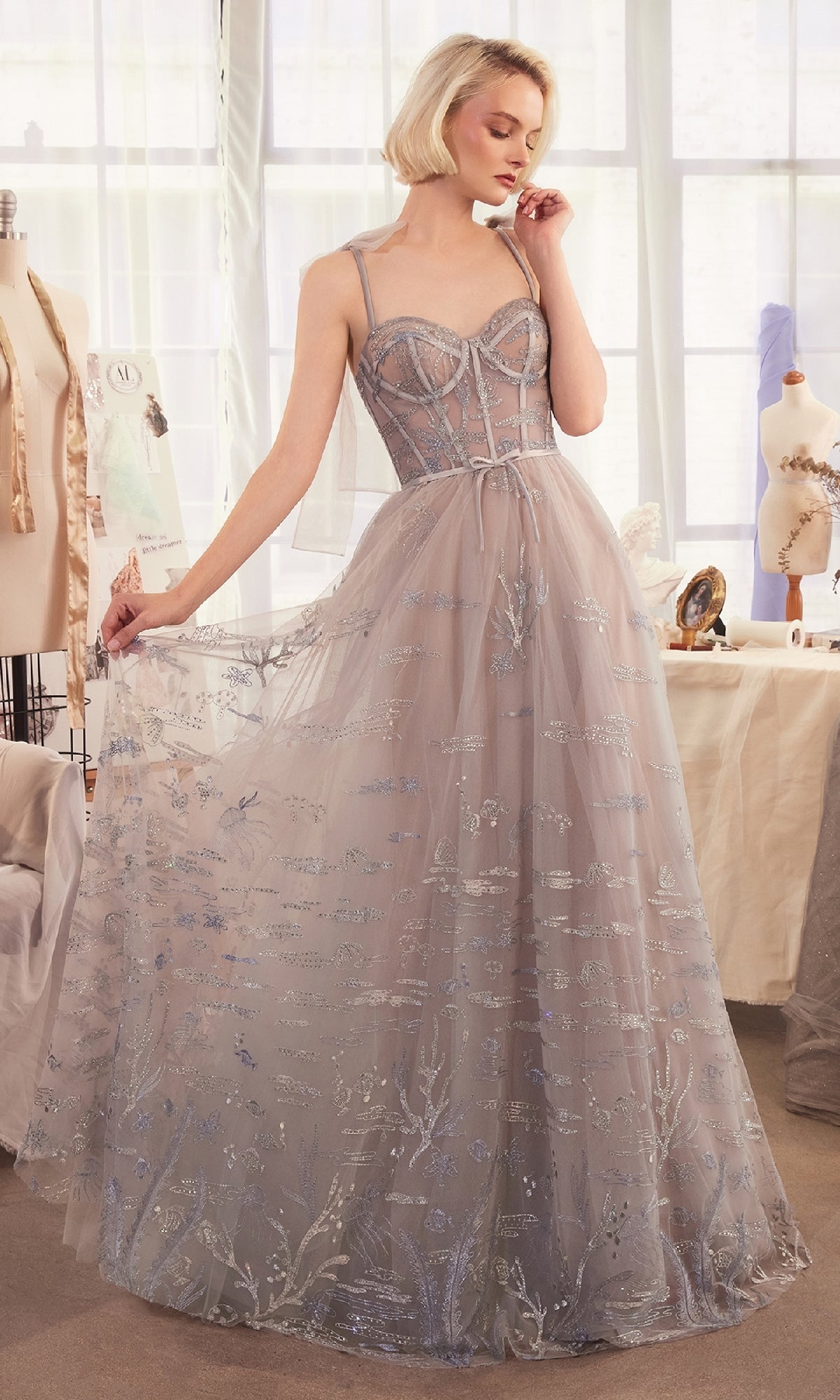 A woman in a Long Silver Prom Ball Gown (Andrea & Leo A1458) stands in a sunlit room with sewing mannequins and dressmaking supplies.