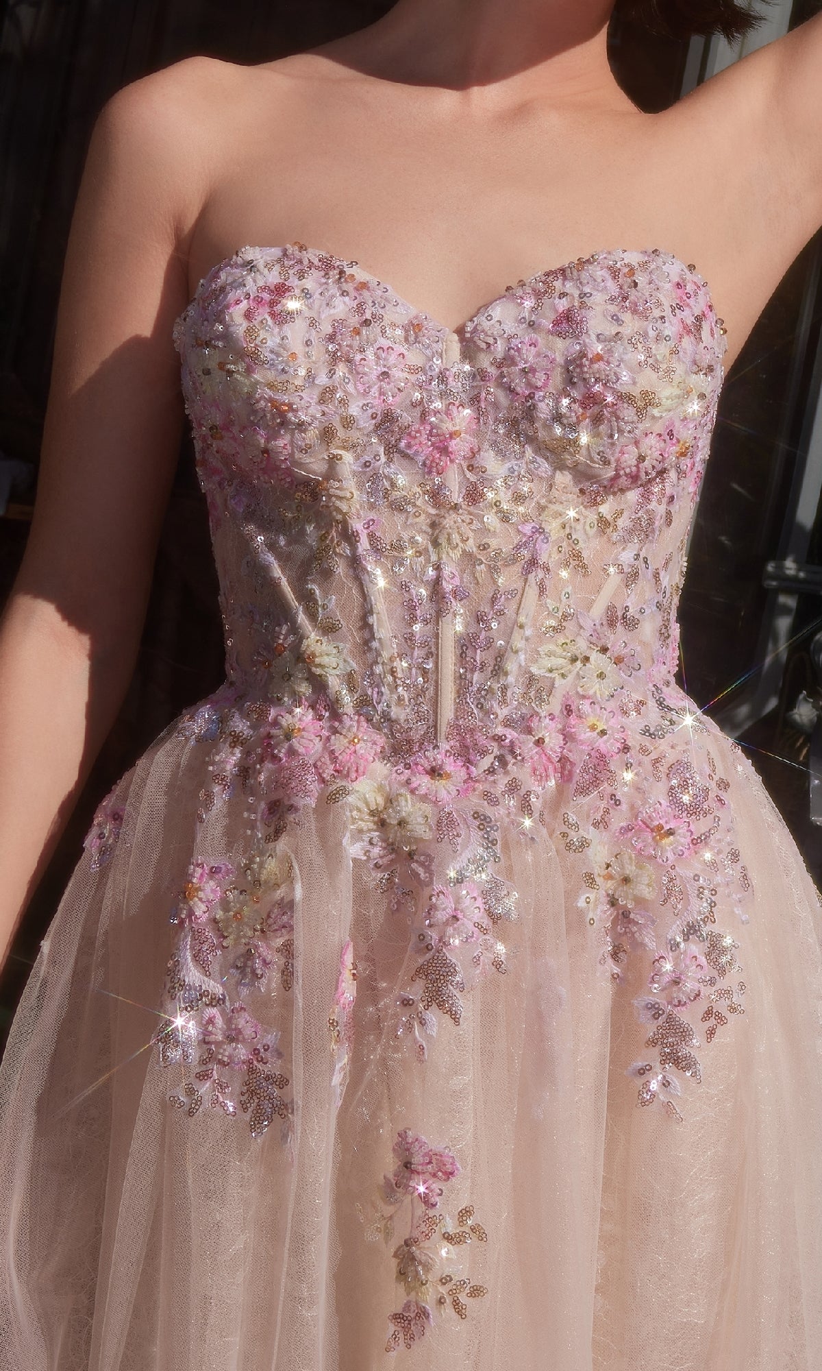 A woman wearing the Strapless Long Prom Ball Gown: Andrea & Leo A1468, featuring a sheer design with pink, white, and purple floral embroidery and sparkling sequins.