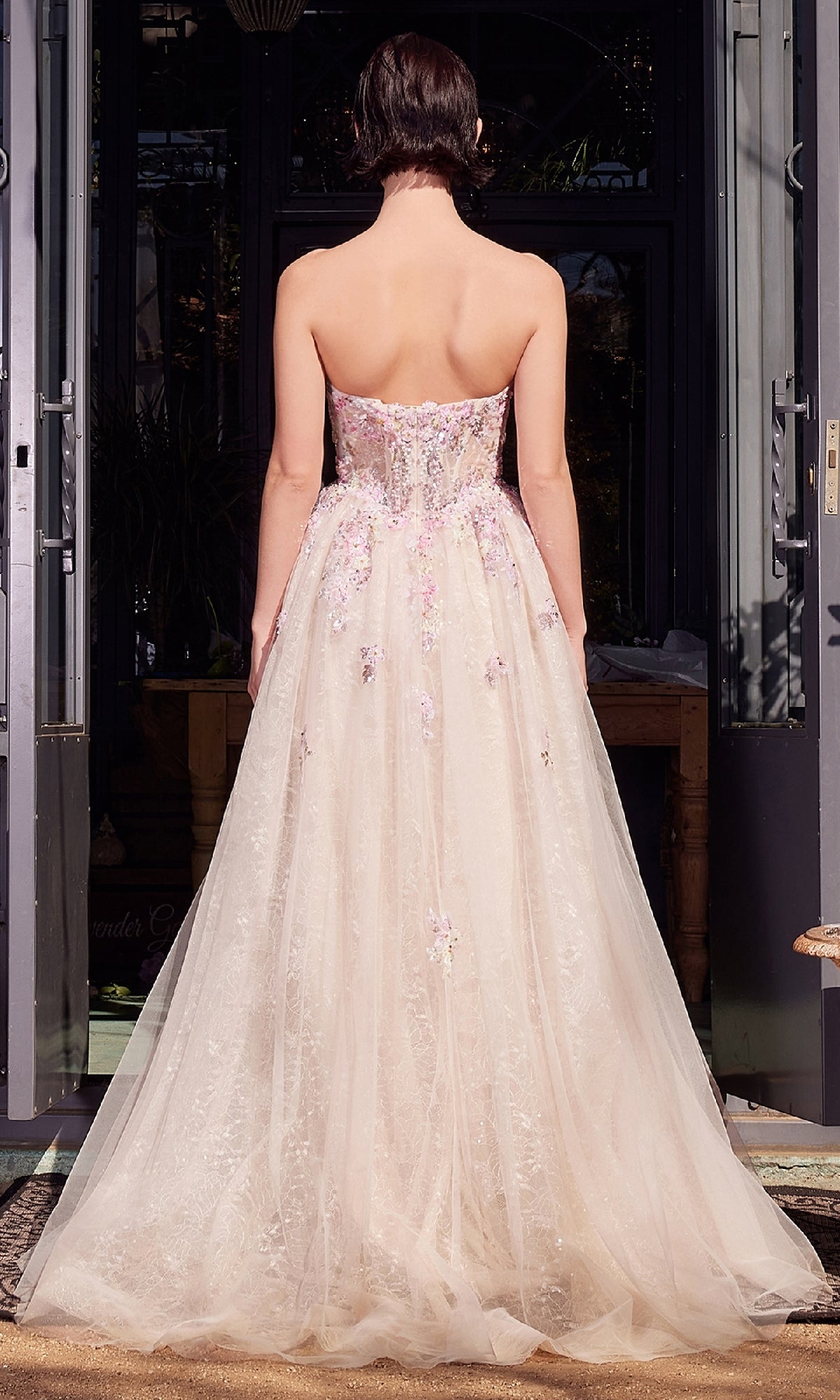 A woman faces away from the camera, wearing the Andrea & Leo A1468 Strapless Long Prom Ball Gown with a sheer embroidered skirt and delicate floral embellishments.