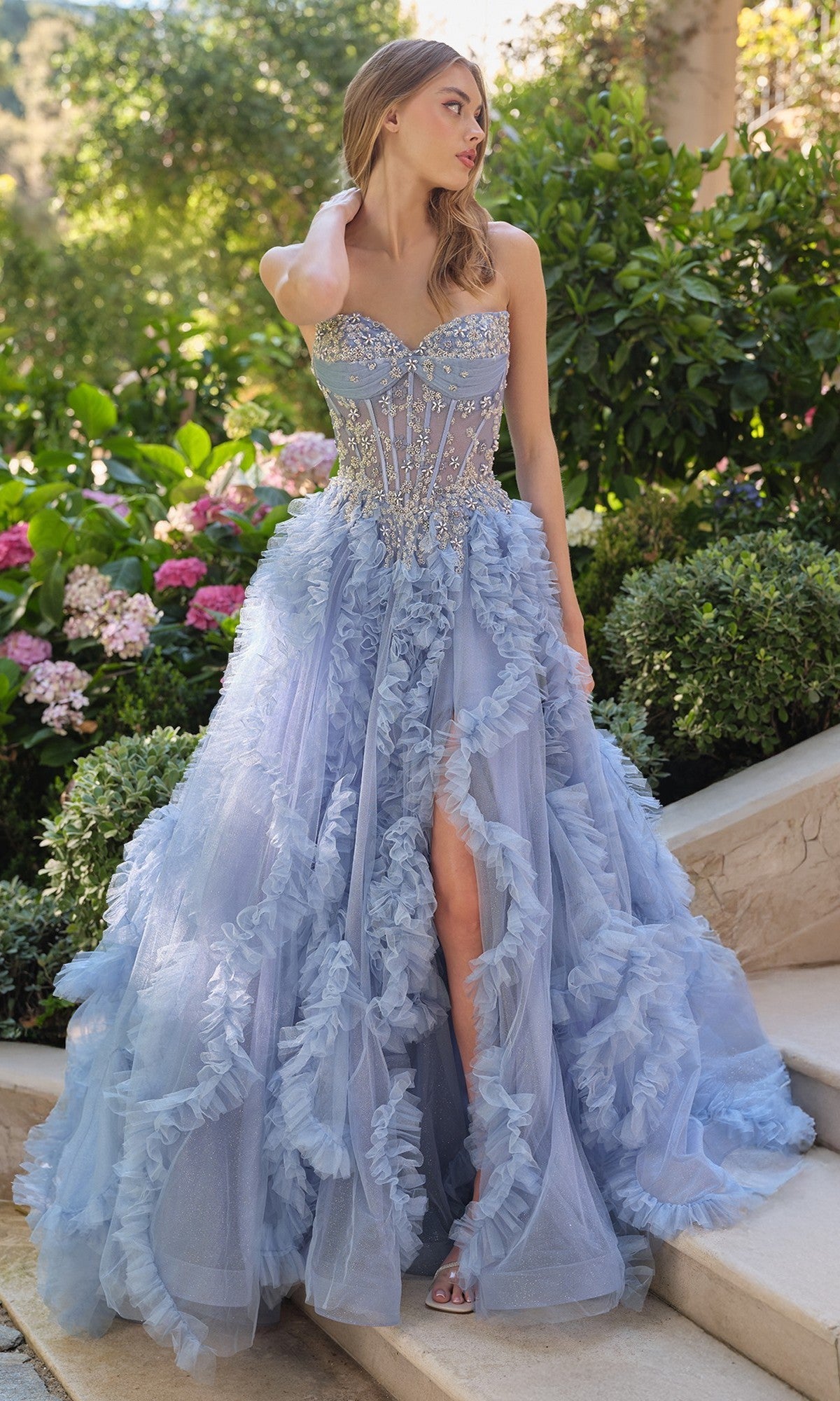 A woman stands outdoors in a Strapless Long Pink Prom Ball Gown, Andrea & Leo A1483, featuring ruffles and surrounded by greenery and flowers.