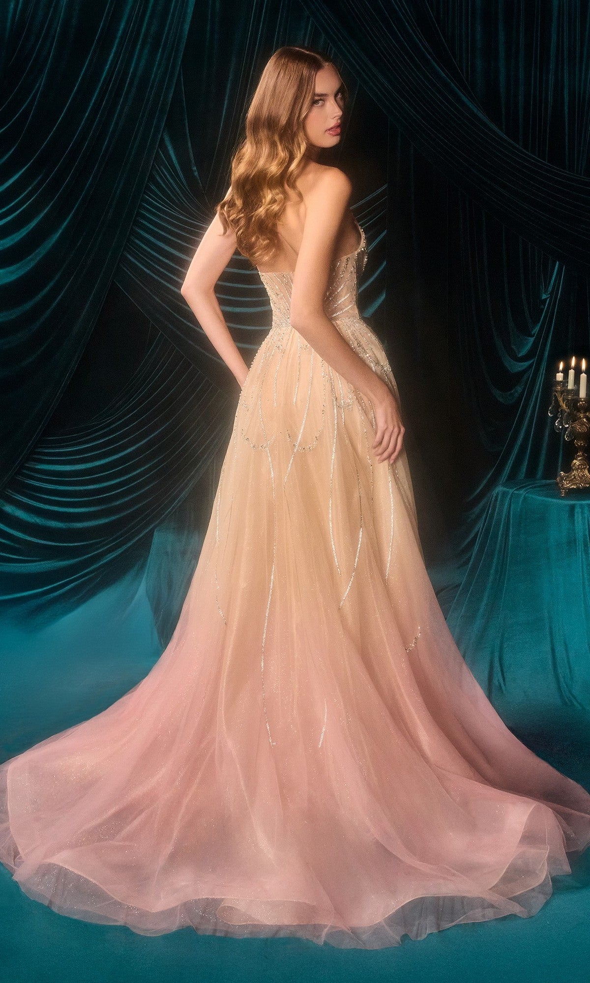 A woman wearing the Andrea and Leo A1508 long prom dress, featuring a strapless peach ombre tulle gown with a flowing train, stands before dark teal drapes near a table with a candelabra.