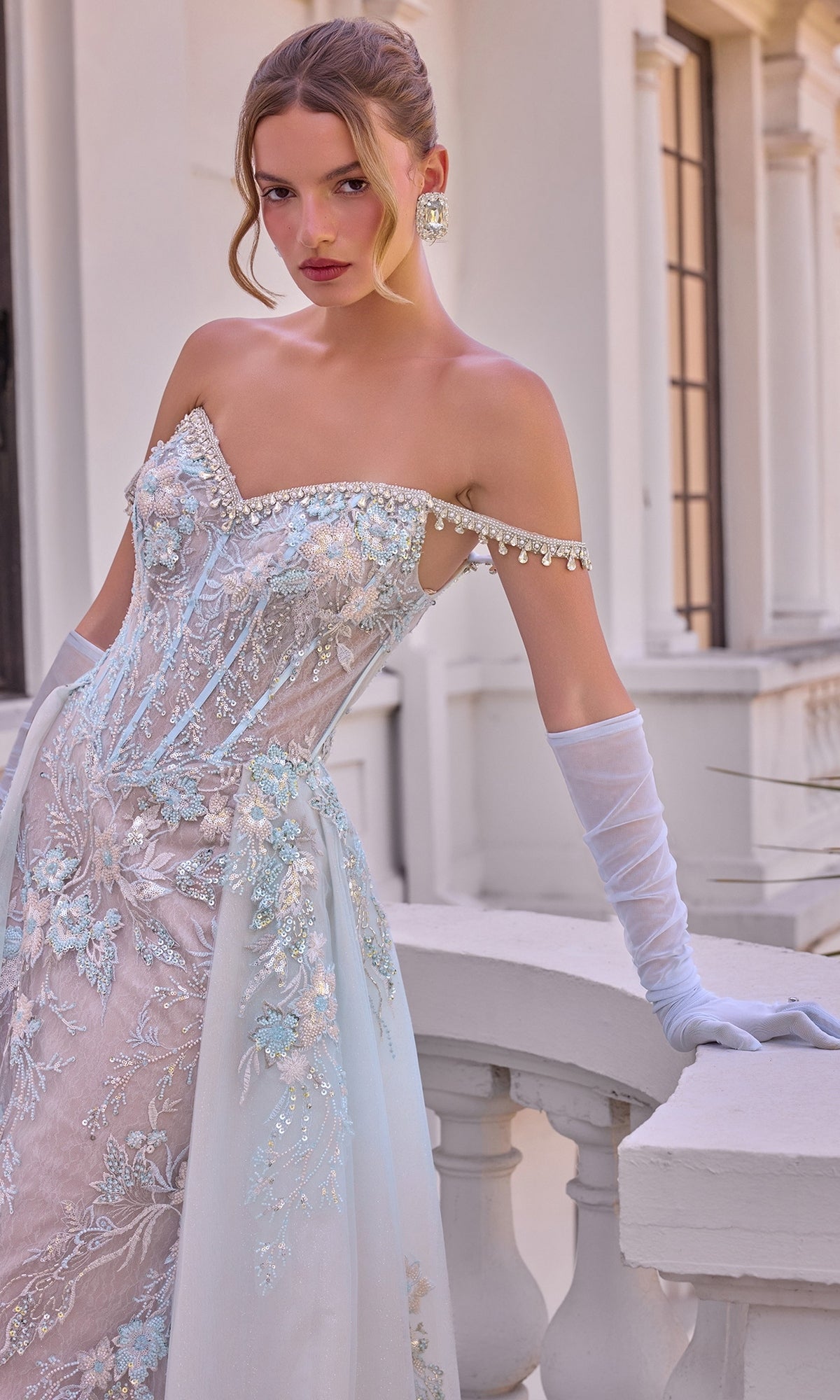 A woman wears the Ethereal Blue Prom Dress: Andrea & Leo A1518, a light blue, beaded, off-the-shoulder corset gown with floral embroidery, sequins, and matching sheer opera gloves.