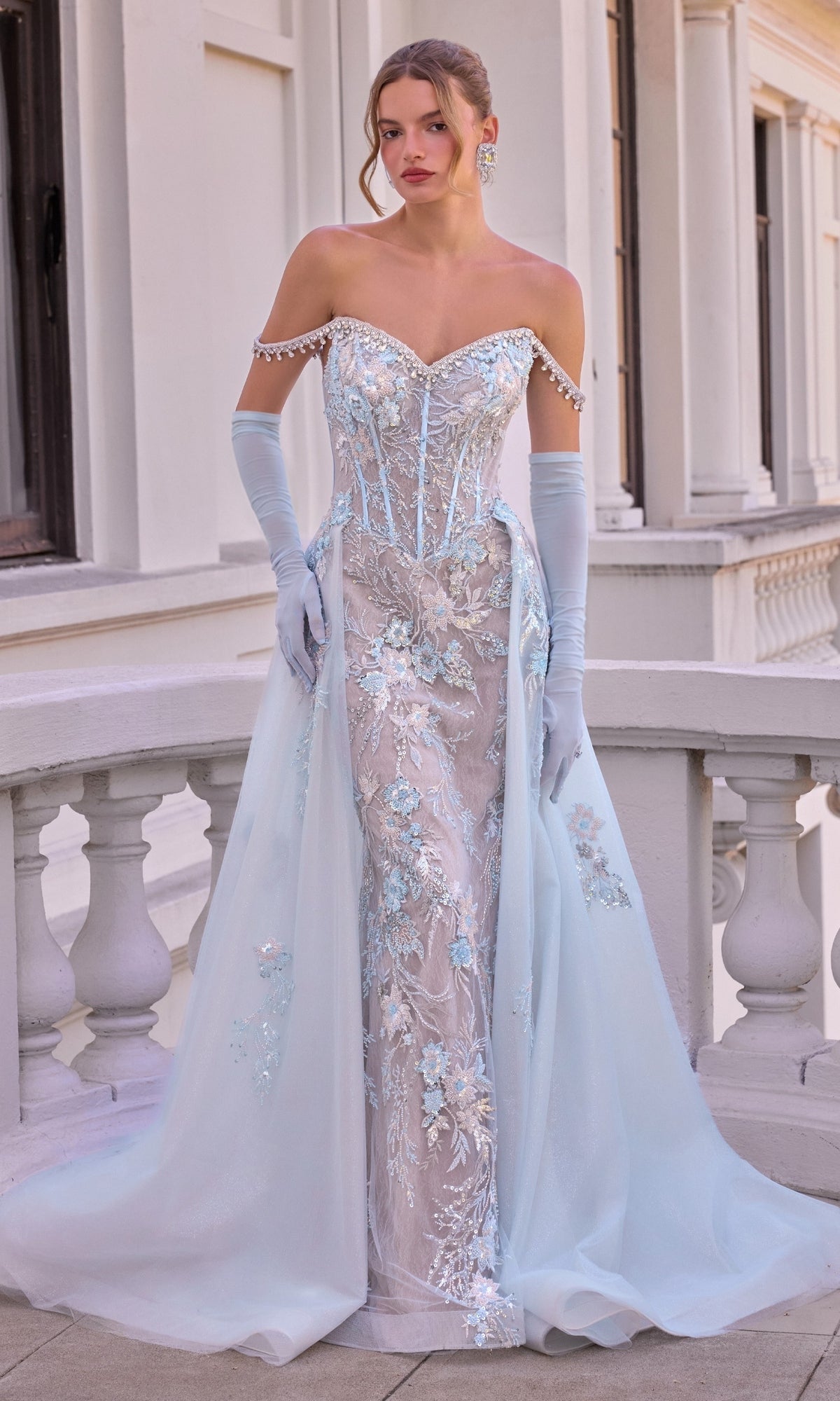 A person wears an Ethereal Blue Prom Dress: Andrea & Leo A1518, an off-the-shoulder blue gown with a corset bodice, sheer overlay, floral embroidery, and matching opera gloves.