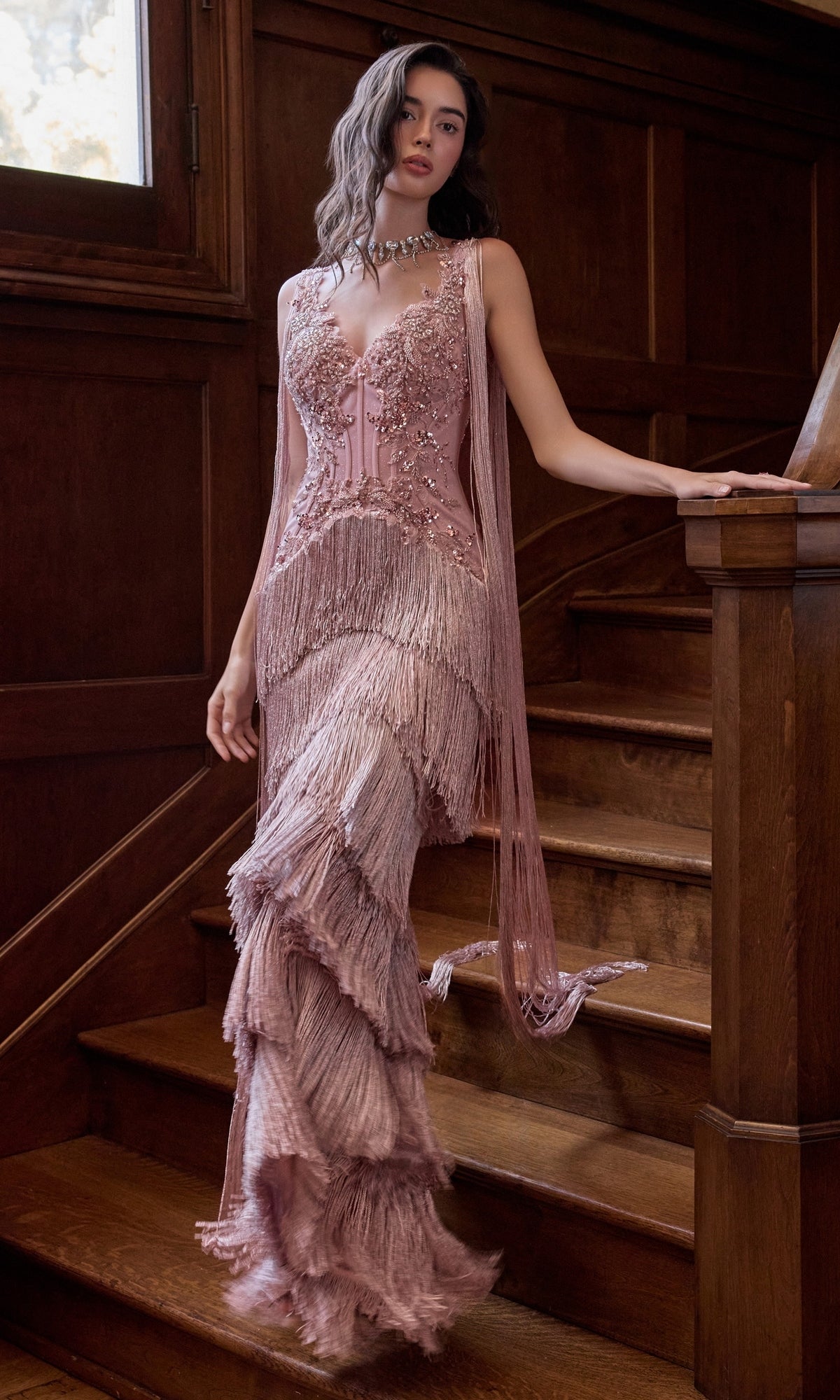 A woman stands on a wooden staircase wearing a mauve-colored Vintage-Inspired Prom Dress: Andrea & Leo A1529, featuring a beaded bodice, fringe detailing, and a layered floor-length skirt.