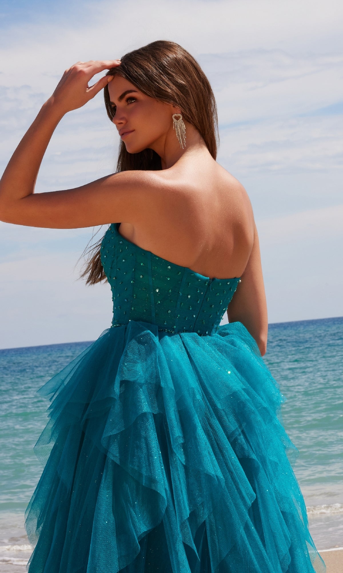 A woman in the Nox Anabel A1591 long prom dress, featuring a strapless corset bodice and ruffled A-line skirt, stands on the beach facing the ocean with one hand gracefully raised to her head.