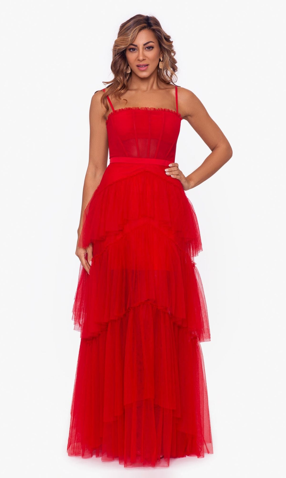 A woman stands facing forward in the Betsy and Adam Long Ruffled Prom Dress A24201, a sleeveless red tulle evening gown with spaghetti straps and layered ruffles, set against a plain white background.