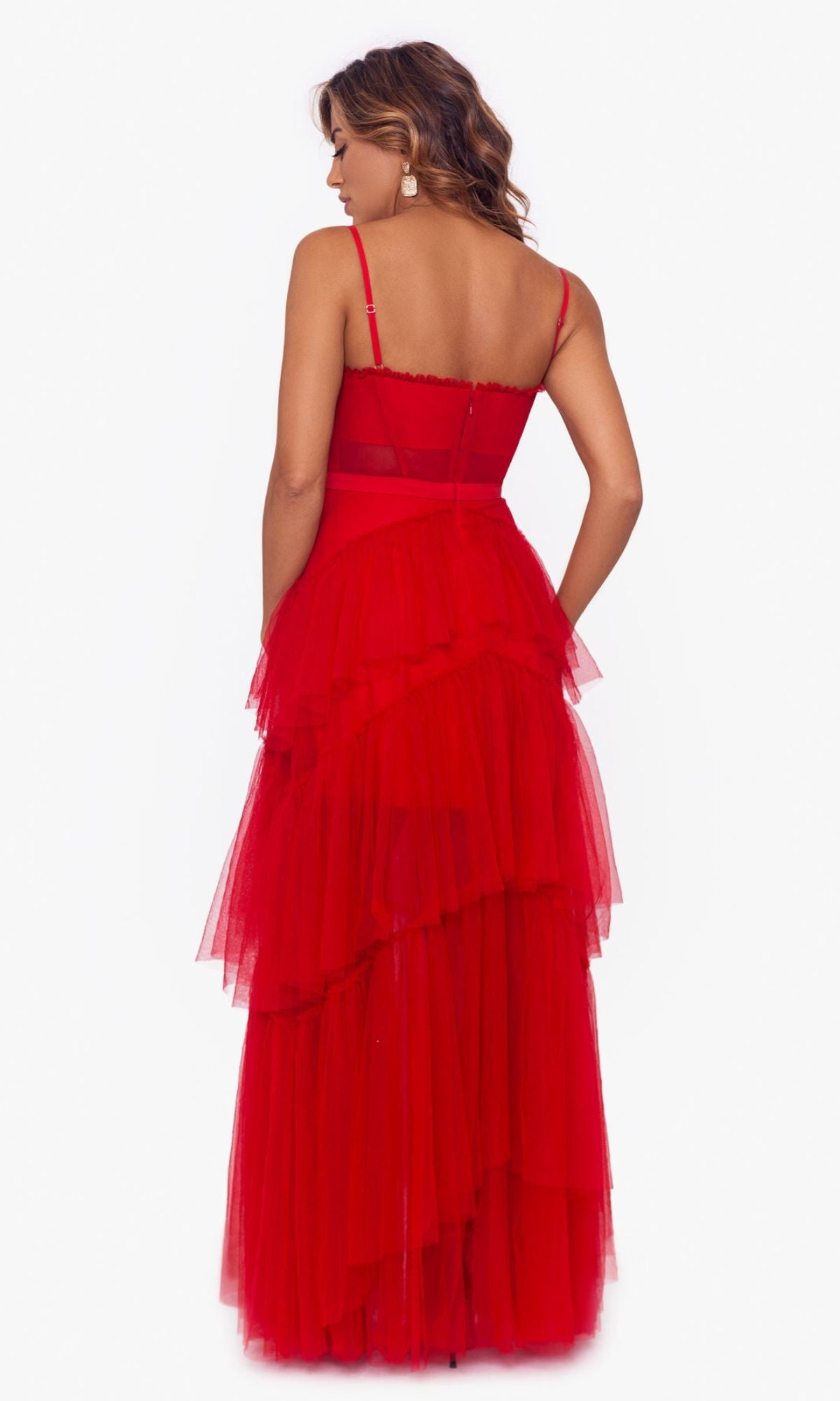 A woman faces away in the Betsy and Adam Long Ruffled Prom Dress A24201, a sleeveless, floor-length tulle gown with tiered ruffles and spaghetti straps, set against a plain white background.