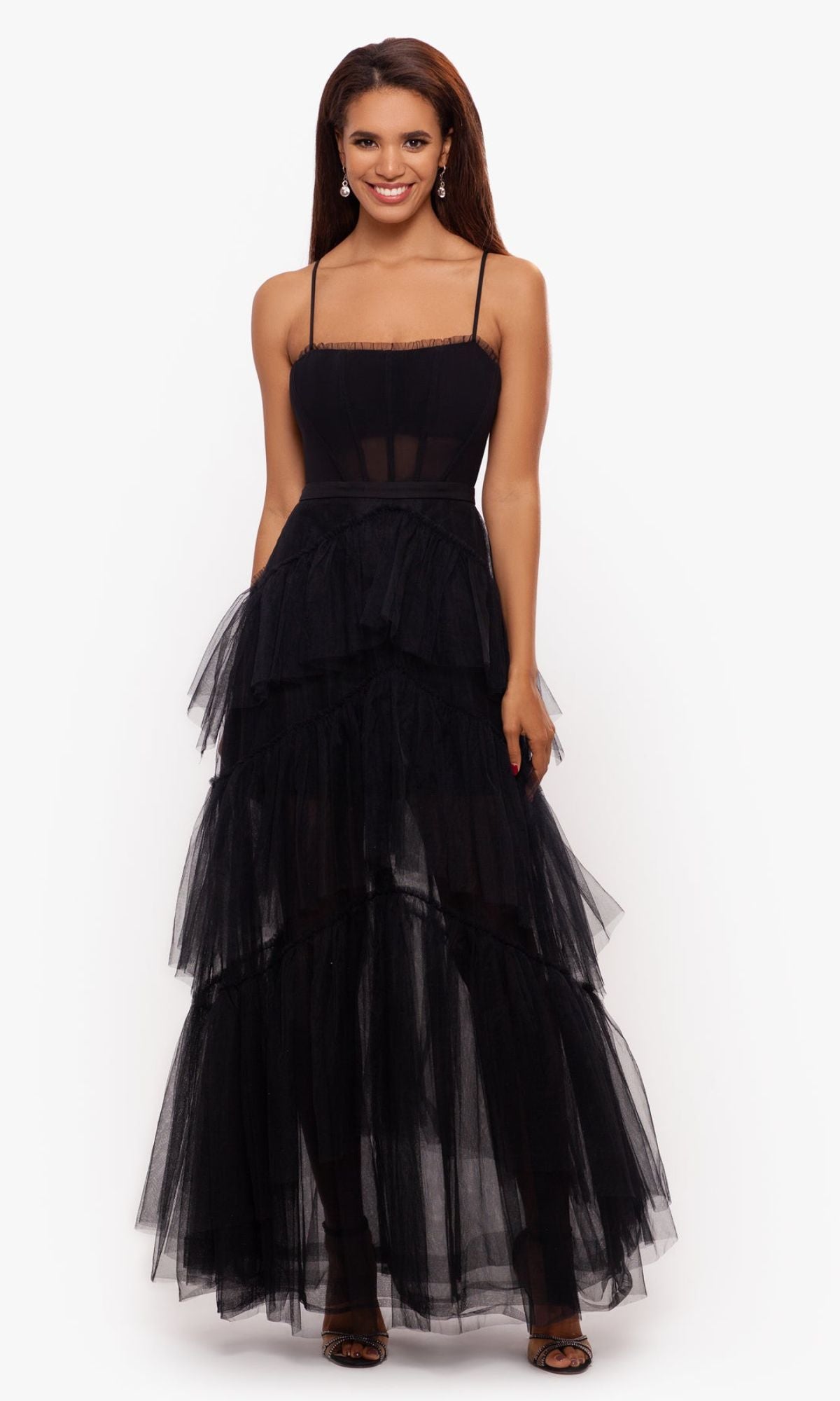 A woman stands smiling in the Betsy and Adam Long Ruffled Prom Dress A24201—a sleeveless black tulle gown with thin straps, sheer bodice, tiered skirt—and black open-toe heels, set against a plain white background.