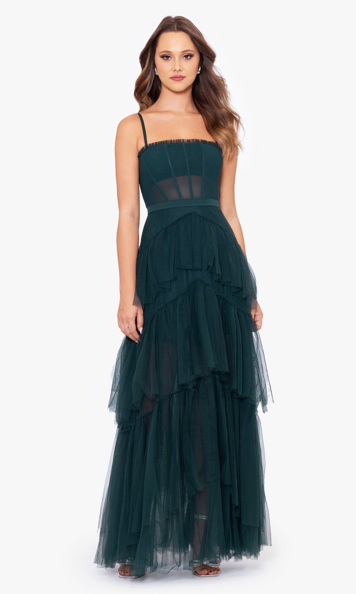 A woman wears the Betsy and Adam Long Ruffled Prom Dress A24201, a dark green sleeveless tulle gown with spaghetti straps and a tiered silhouette, standing against a plain white background.