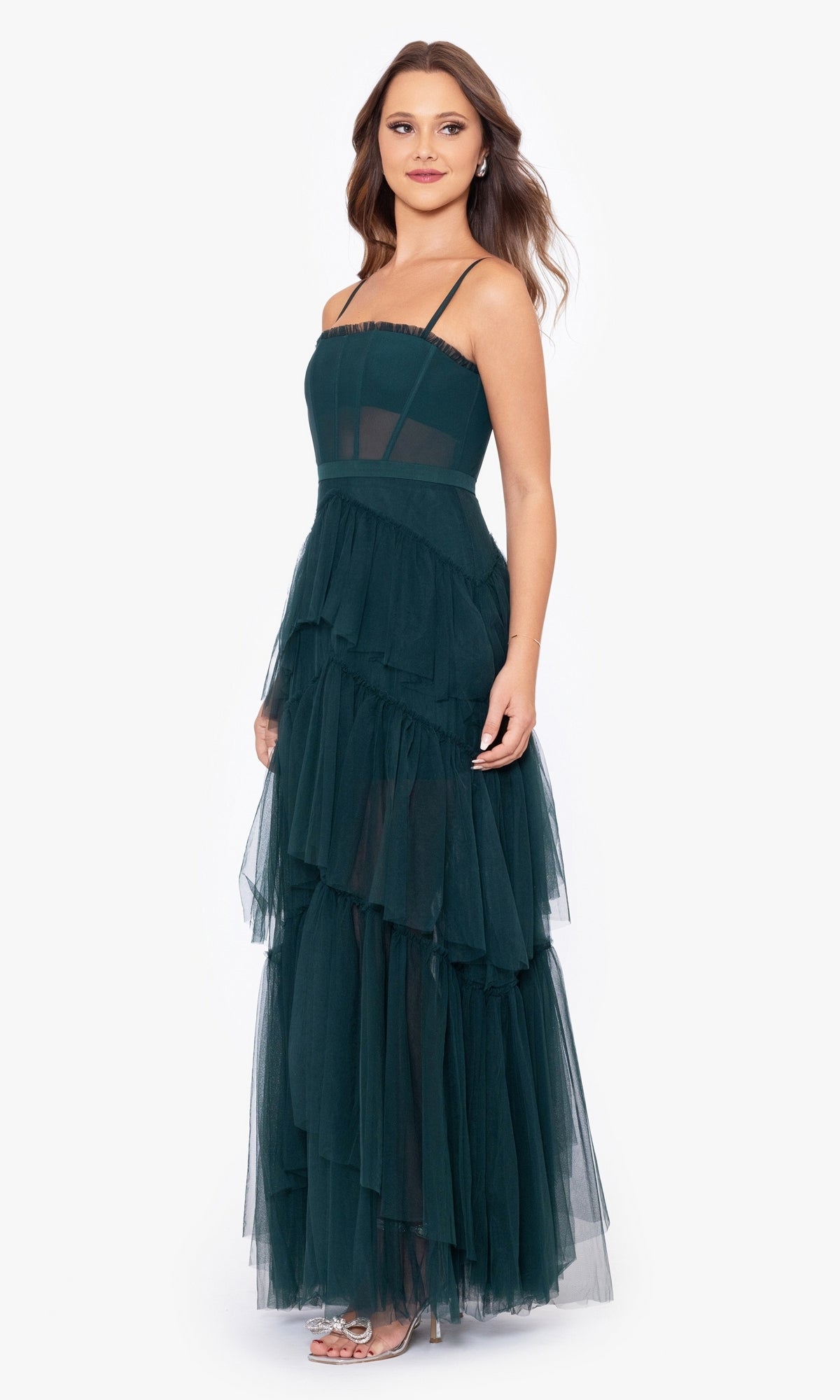 A woman stands against a plain white background wearing the Betsy and Adam Long Ruffled Prom Dress A24201 in dark green, featuring spaghetti straps, sheer paneling, and tiered tulle layers for an elegant evening look.