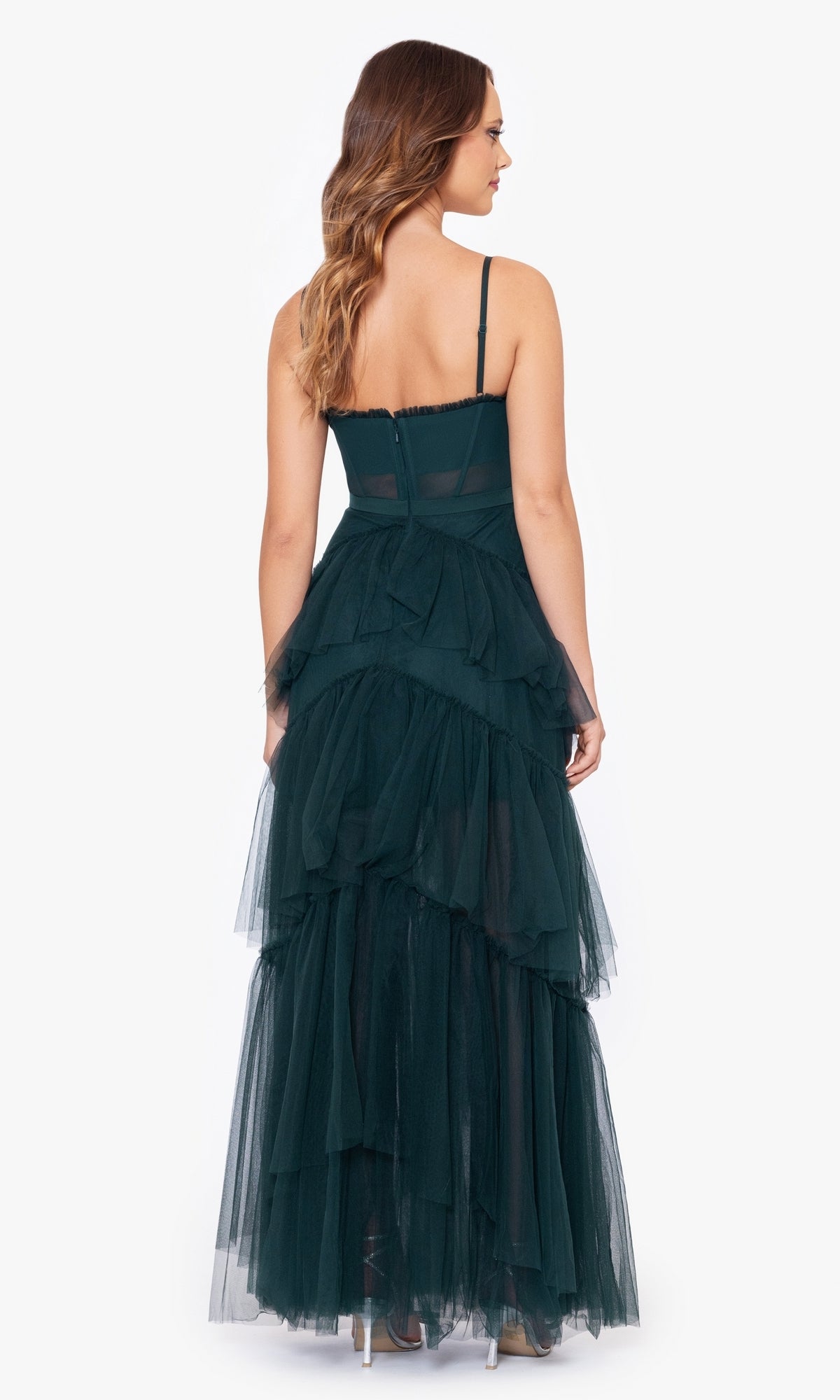 A woman stands with her back to the camera, wearing the Betsy and Adam Long Ruffled Prom Dress A24201 in dark green, featuring thin straps and tiered layers, styled with high heels against a plain white background.