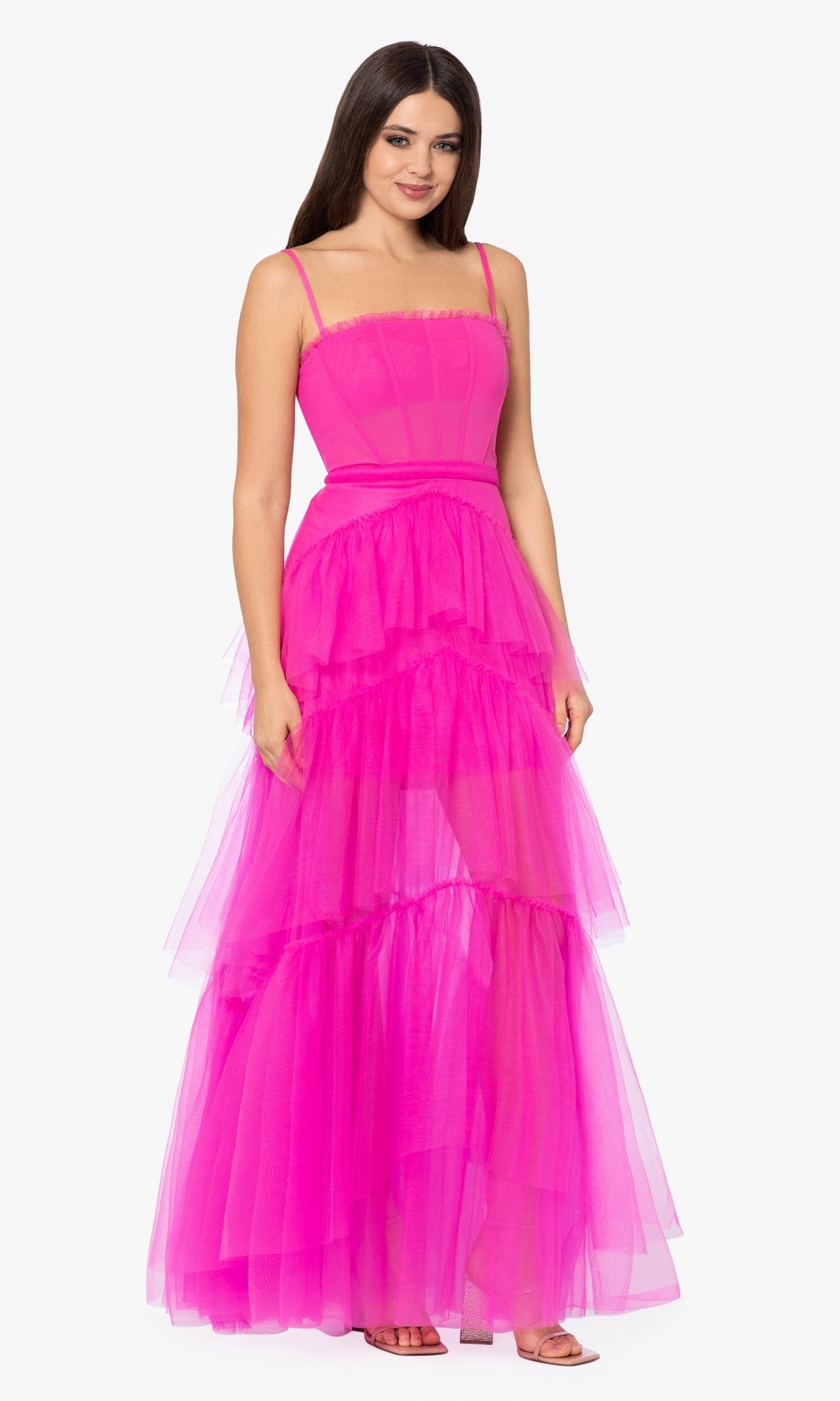 A woman models the Betsy and Adam Long Ruffled Prom Dress A24201 in bright pink with spaghetti straps, standing against a plain white background.