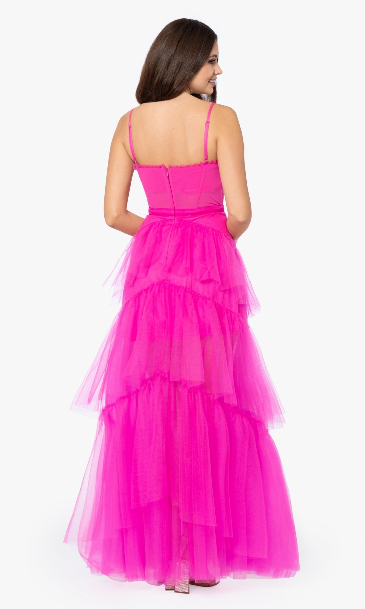 A woman is shown from the back wearing the Betsy and Adam Long Ruffled Prom Dress A24201 in bright pink, featuring tiered tulle, against a plain white background.