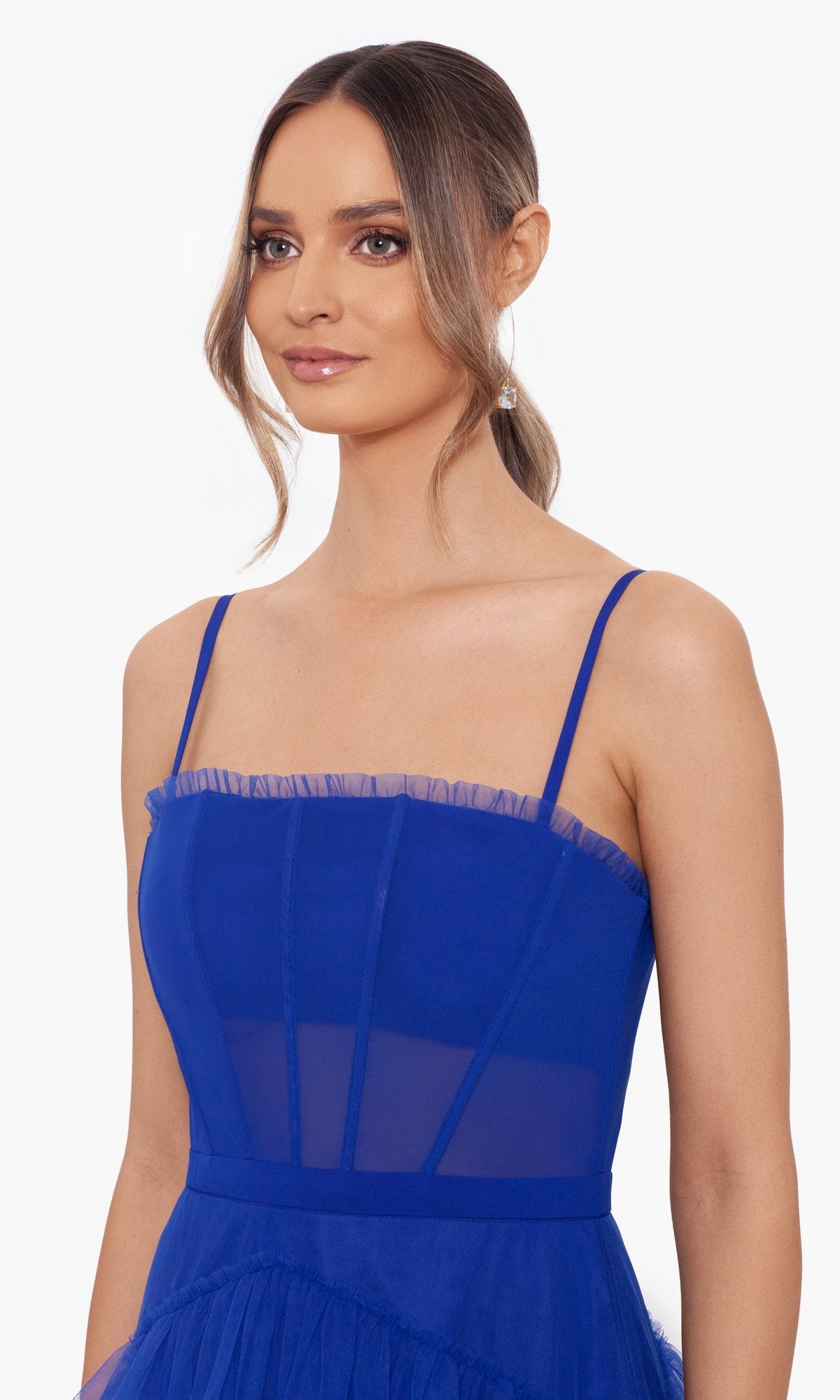 A woman with long brown hair in a low ponytail wears the Betsy and Adam Long Ruffled Prom Dress A24201, a royal blue formal gown with thin straps and tiered sheer details, standing against a plain white background.
