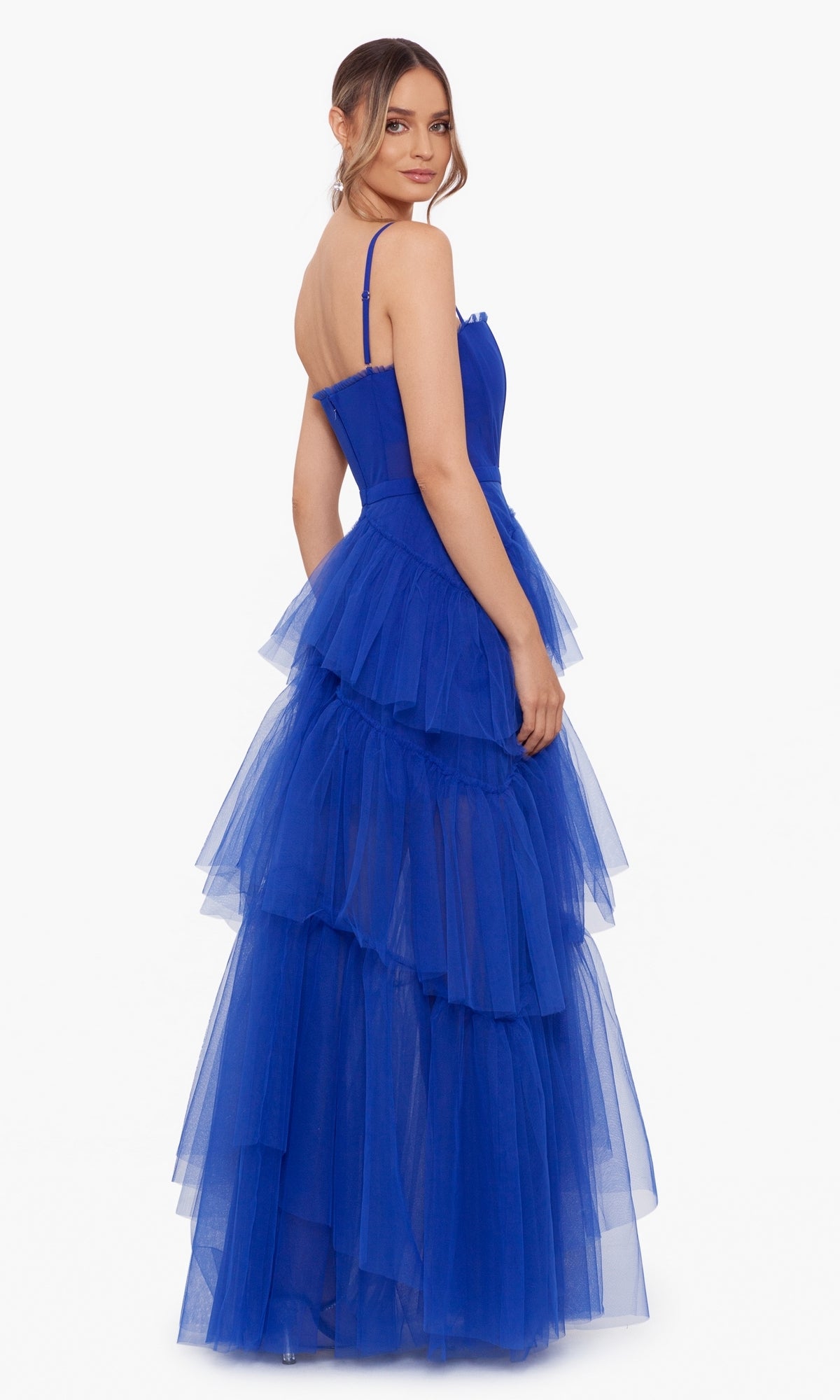 A woman in the Betsy and Adam Long Ruffled Prom Dress A24201, a sleeveless royal blue evening gown with tiered tulle, poses slightly turned away, her hair styled in a low ponytail.