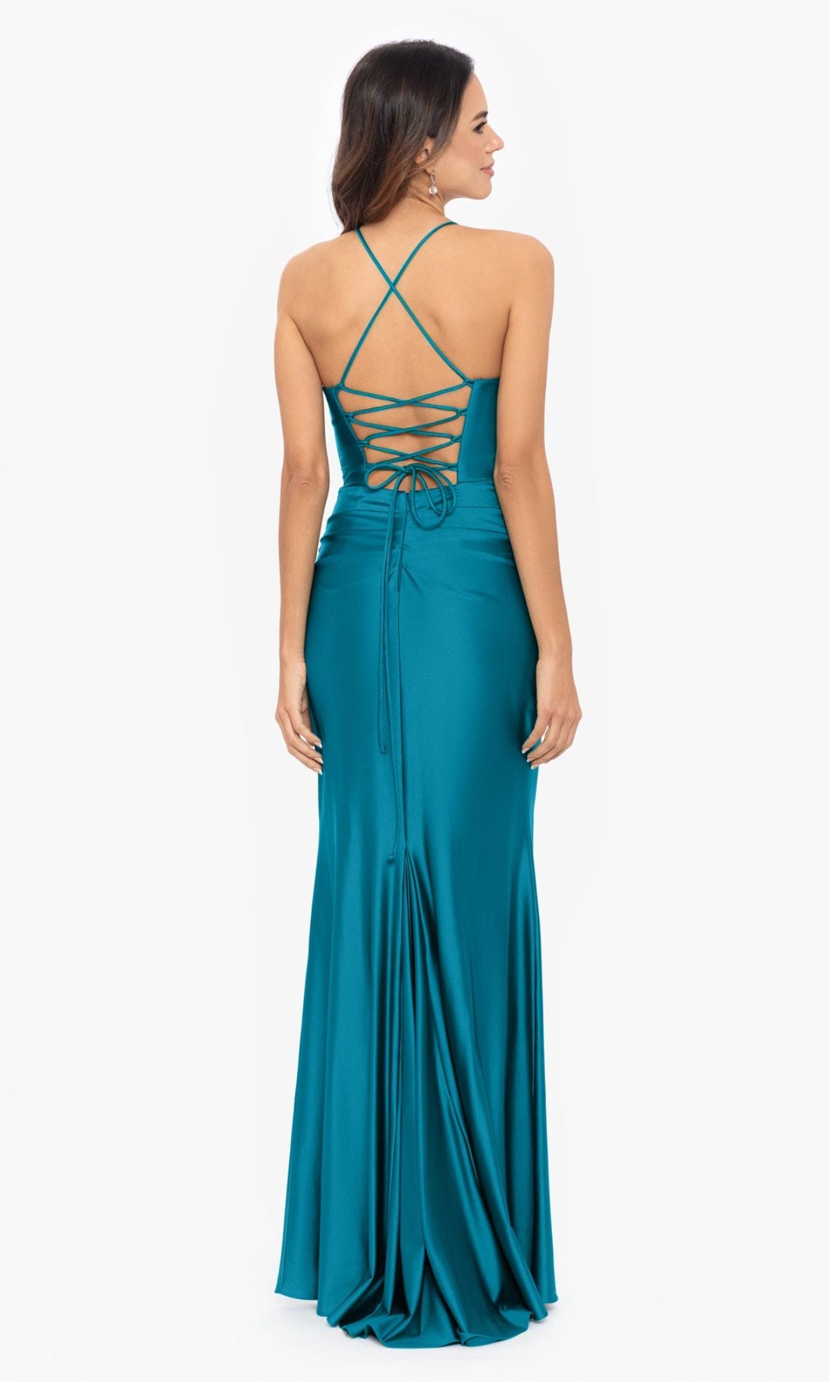 A woman stands facing away from the camera in the Betsy and Adam Long Green Prom Dress A25610, featuring a corset-style bodice, crisscross lace-up back, and a floor-length teal hem.