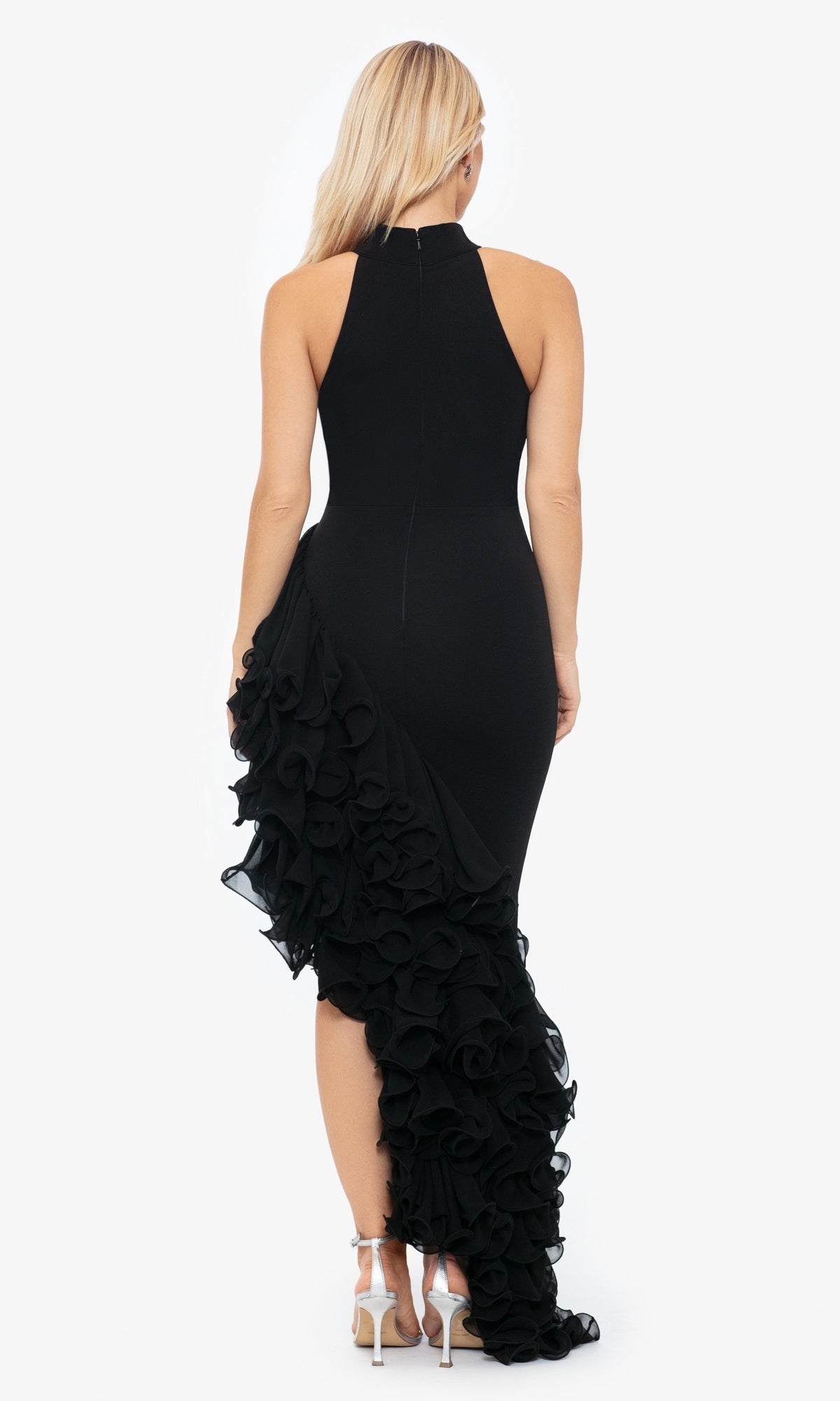 Woman wearing the Betsy and Adam Long Black Ruffled Prom Dress A26173, featuring a sleeveless high neckline and ruffled side detail, paired with chic high-heeled sandals and posing with her back to the camera.