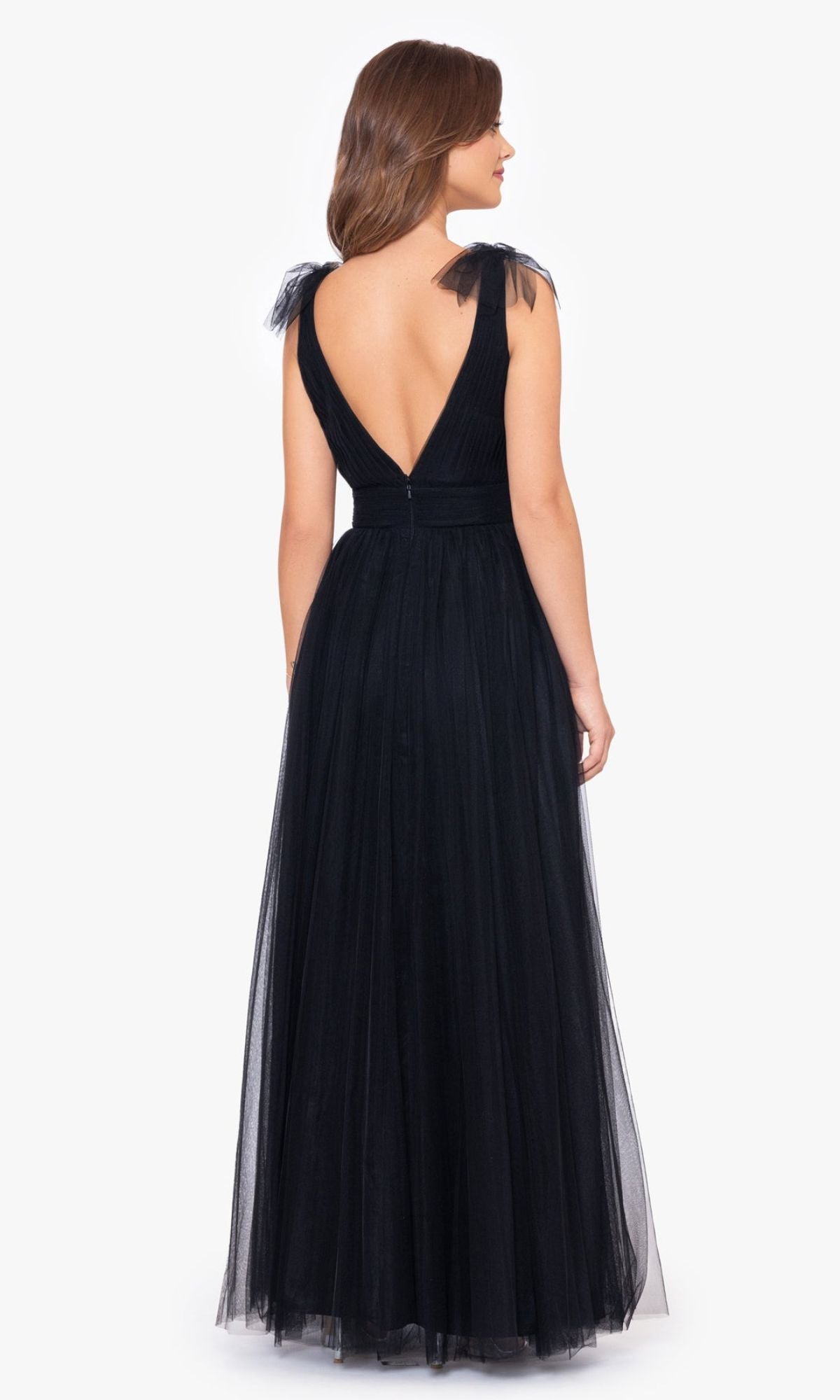 A woman faces away in the Betsy & Adam Shoulder-Bow Long Prom Dress A26256—a floor-length black tulle gown with a deep V-back and sheer shoulder details—posed against a plain white background.