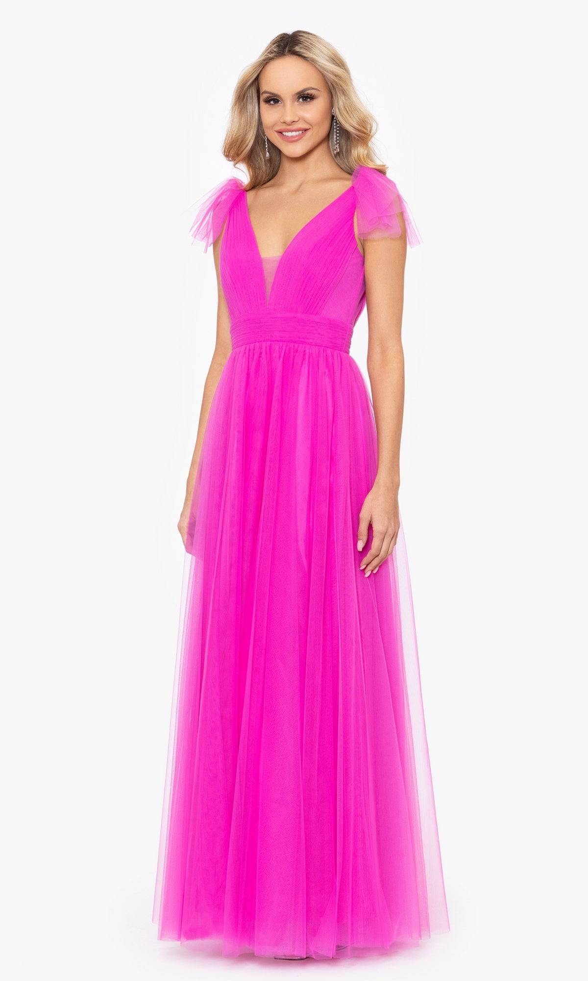 A woman stands and smiles against a white background, wearing the Betsy & Adam Shoulder-Bow Long Prom Dress A26256— a floor-length bright pink gown with cap sleeves, a deep V-neckline, and a fitted waist.
