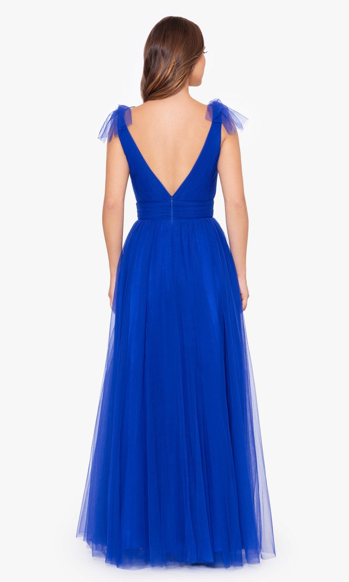 A woman is shown from behind wearing the Betsy & Adam Shoulder-Bow Long Prom Dress A26256, a floor-length deep blue gown with a deep V-back and sheer shoulder straps.