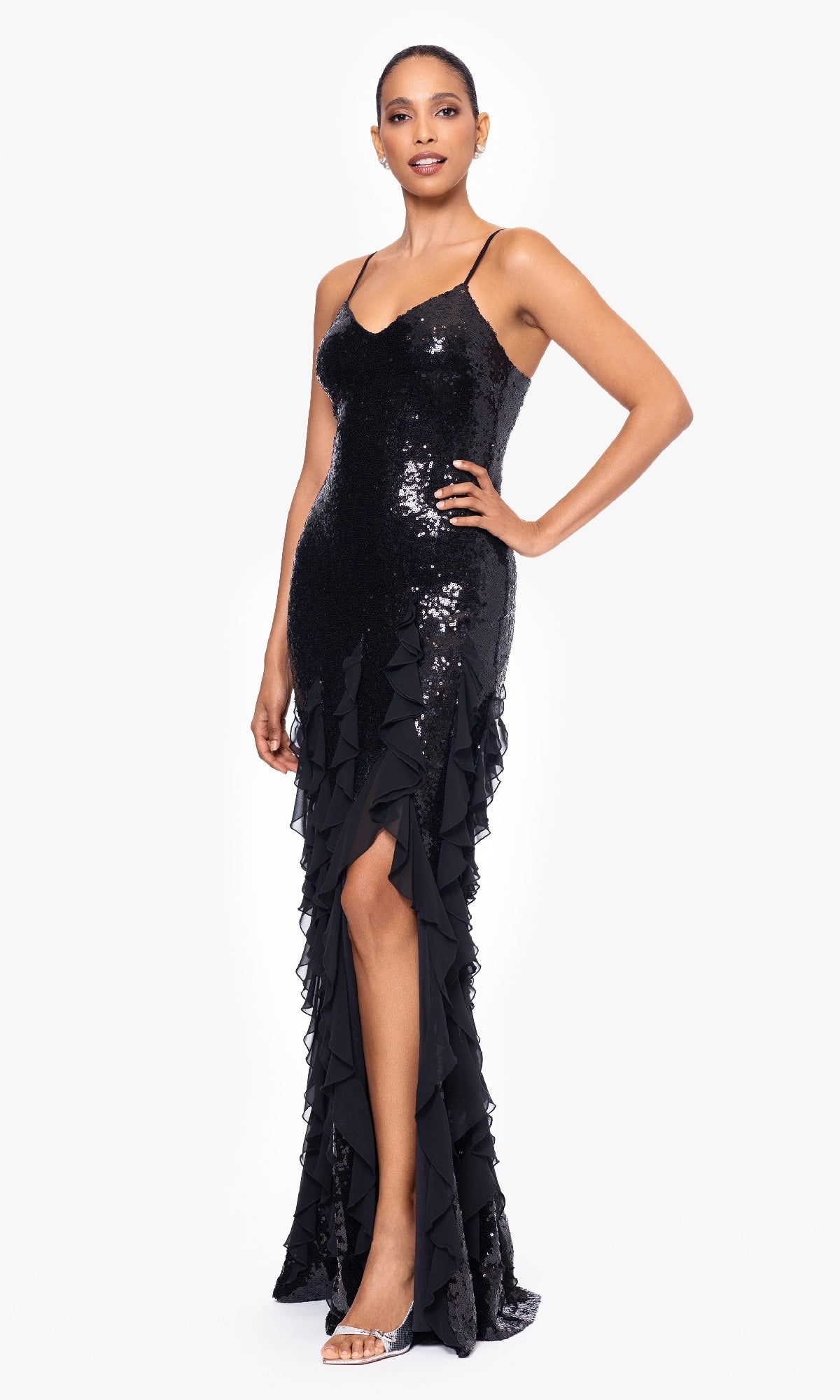 A woman stands wearing the Betsy & Adam A26805: a sleeveless, floor-length black sequin formal gown with ruffle details and a thigh-high slit, paired with silver open-toe heels.