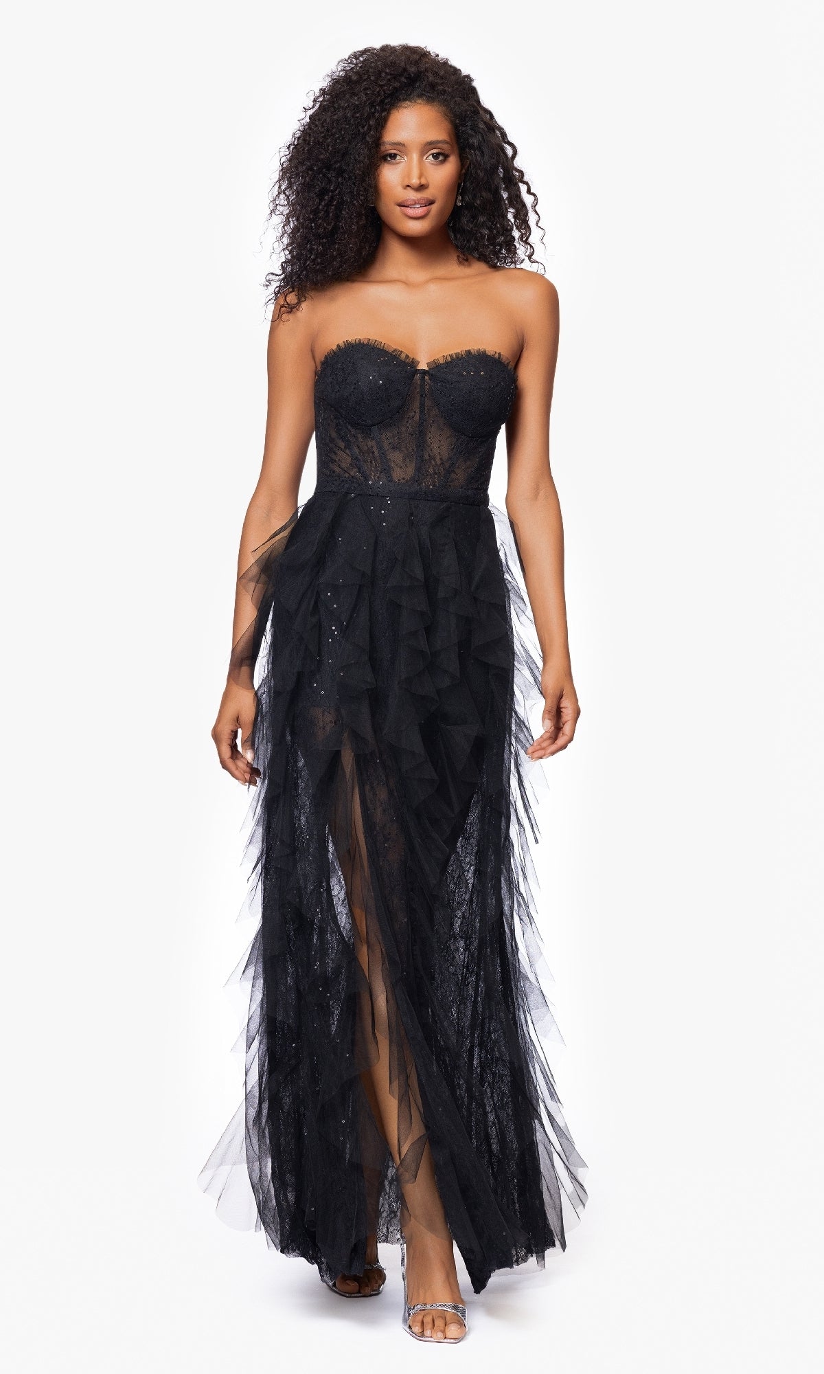 A woman walks forward against a white background, wearing the Betsy & Adam A26845 Strapless Black Prom Gown featuring sheer panels and ruffled tulle accents.