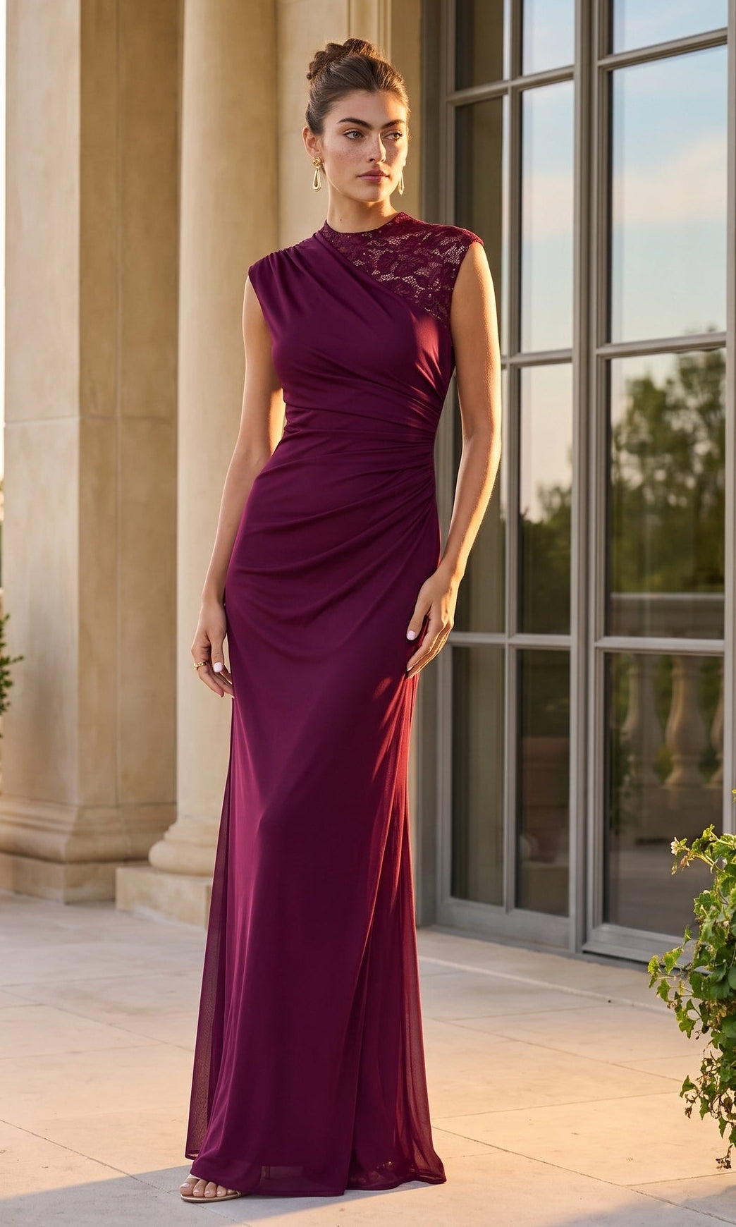 A woman stands outdoors in front of tall windows, wearing the Betsy & Adam A26945 long formal dress in garnet/nude, which features a sleeveless silhouette and delicate lace detailing on one shoulder.