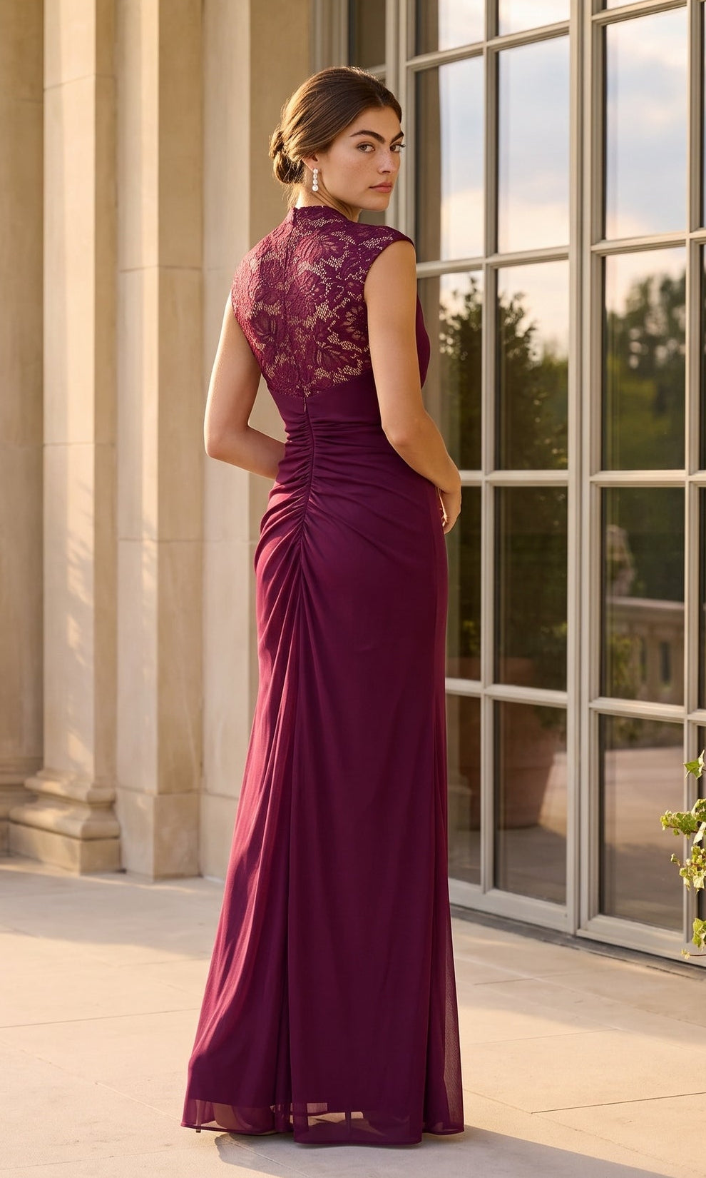 A woman wearing the Long Formal Dress: Betsy & Adam A26945, which features elegant lace detailing on the back, stands outdoors near large windows and looks over her shoulder.