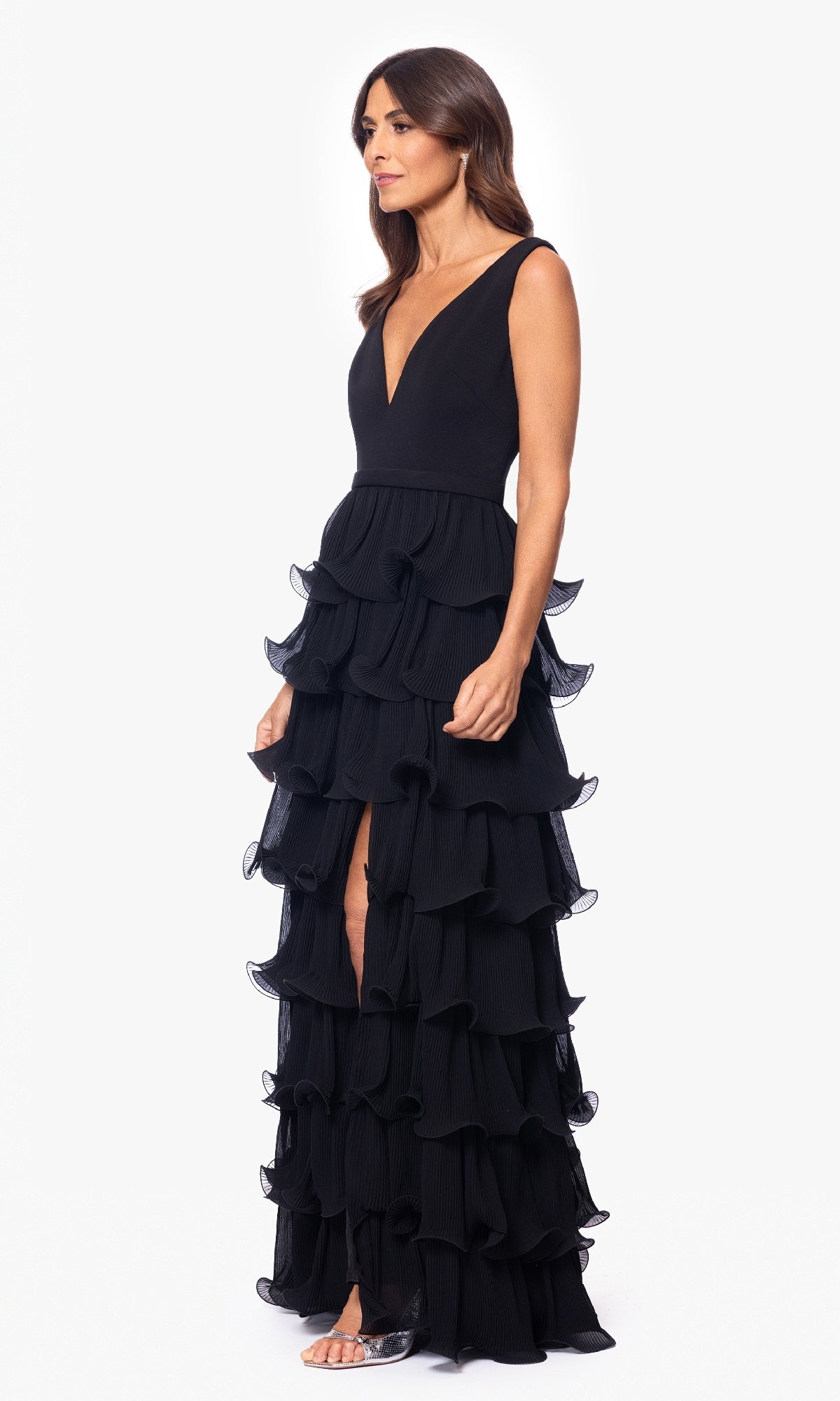 A woman stands against a plain white background wearing the Betsy and Adam A26956 long formal dress—a sleeveless black gown with a deep V-neckline, layered ruffle skirt, front slit—and clear open-toe heels.