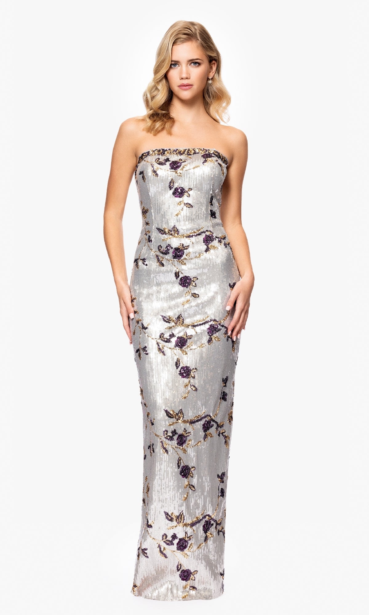 A woman wears the Betsy and Adam A26994 long formal dress, a form-fitting, strapless silver gown with purple and gold floral sequins, standing against a plain white background.
