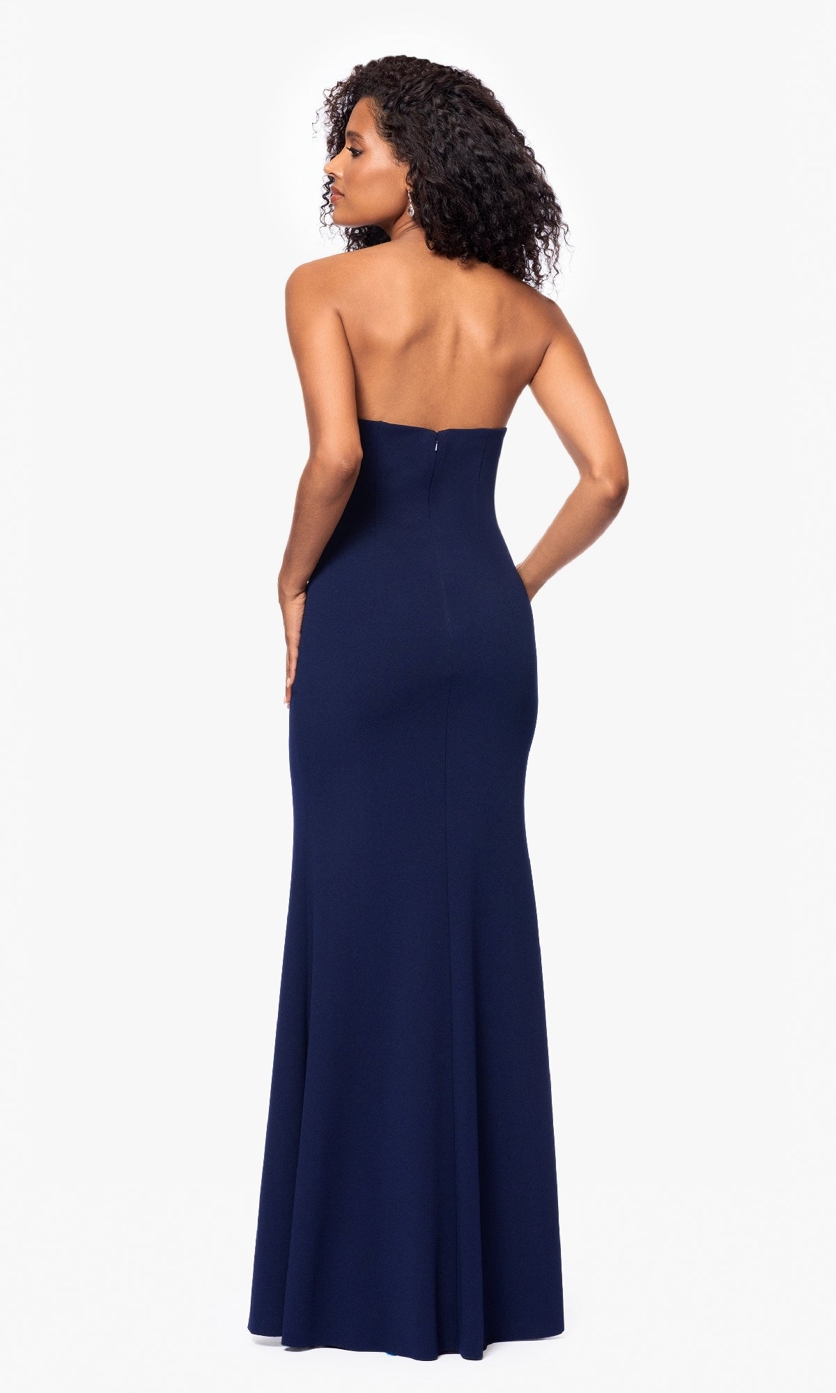 A woman with curly hair models the Betsy and Adam A27030 long formal dress, strapless with a beaded bodice in navy blue, floor-length, shown from the back against a plain white background.
