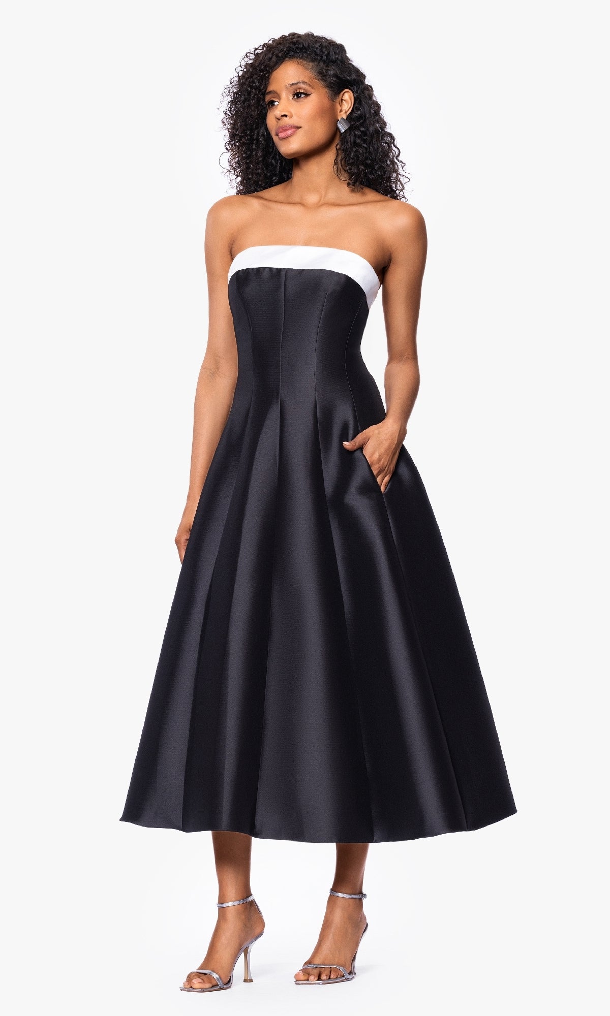 A woman wears the Betsy and Adam A27115 tea-length black formal dress, a strapless A-line midi with a white neckline band, paired with strappy high-heeled sandals.