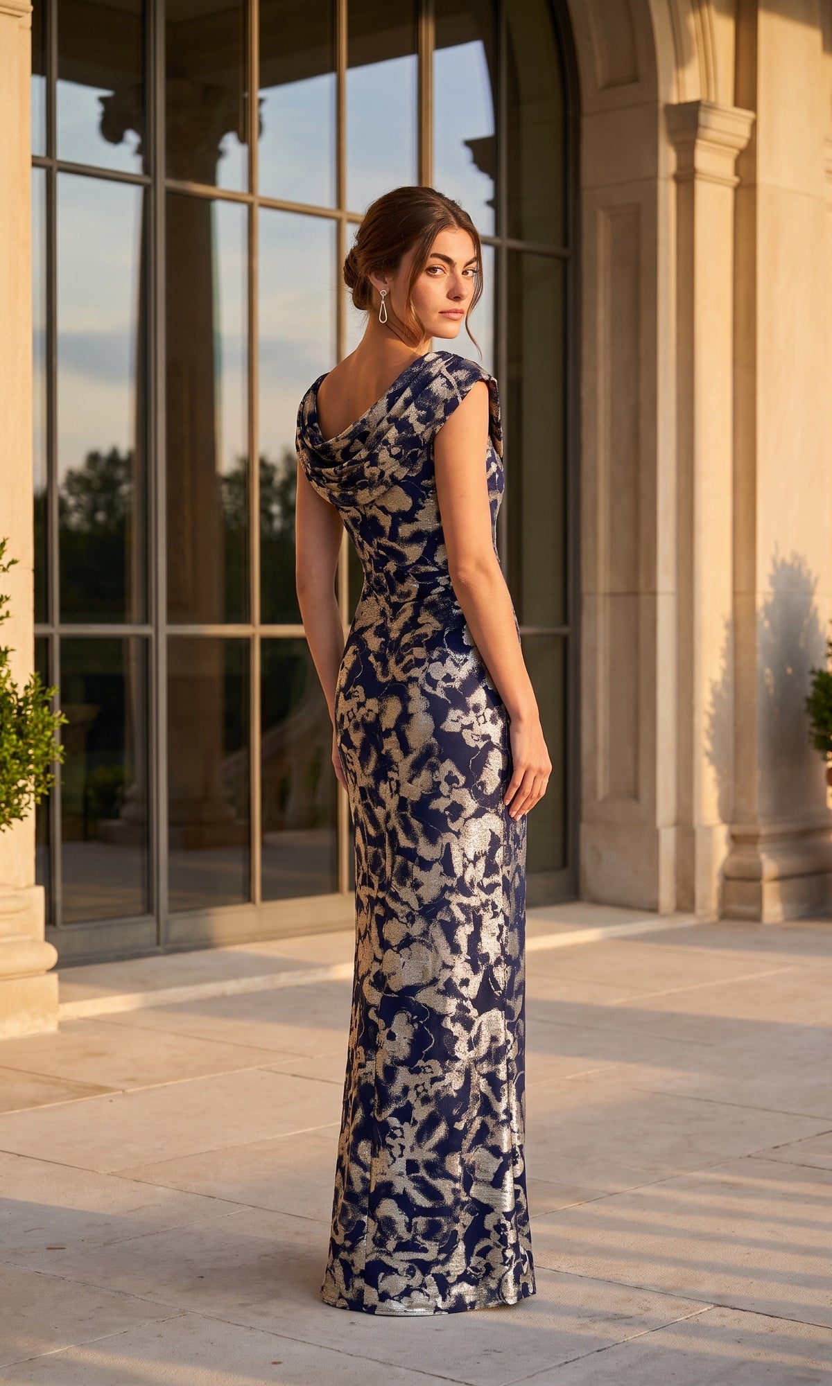 A woman wearing the Betsy & Adam A27195 Long Formal Dress in navy-blue with shimmering gold metallic foil fabric stands outside a building with large windows and columns, looking over her shoulder.