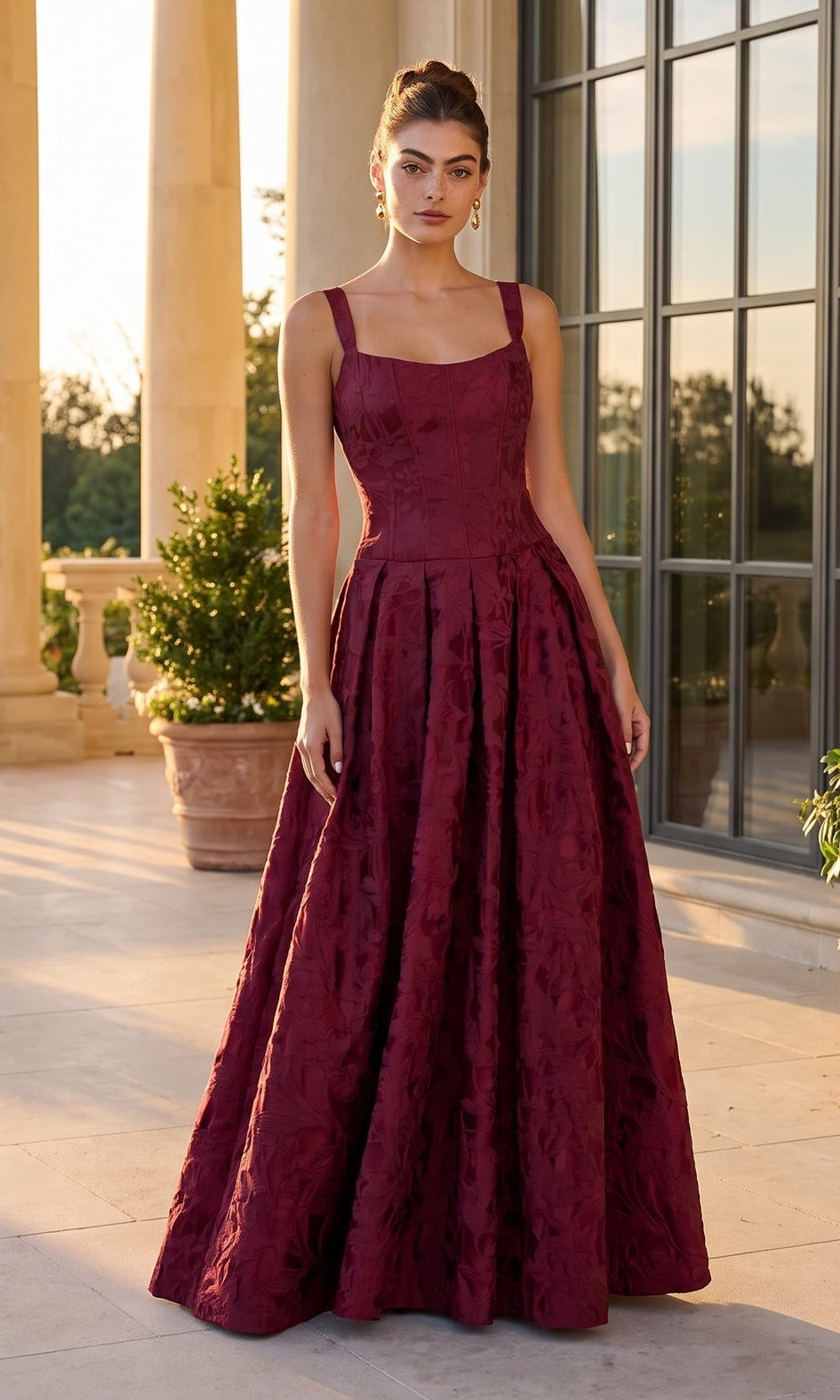 A woman stands on a sunlit terrace with columns and potted plants in the background, wearing the Betsy & Adam A28250 long formal dress in burgundy.