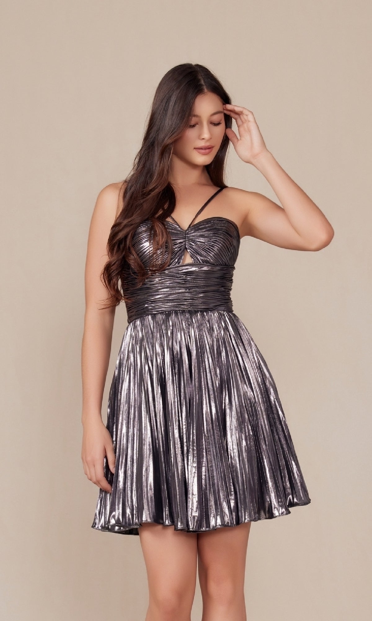 A woman models the Pleated Metallic Homecoming Dress (Nox Anabel A825), strapless and shimmery, against a beige backdrop, one hand in her hair and the other resting at her side.