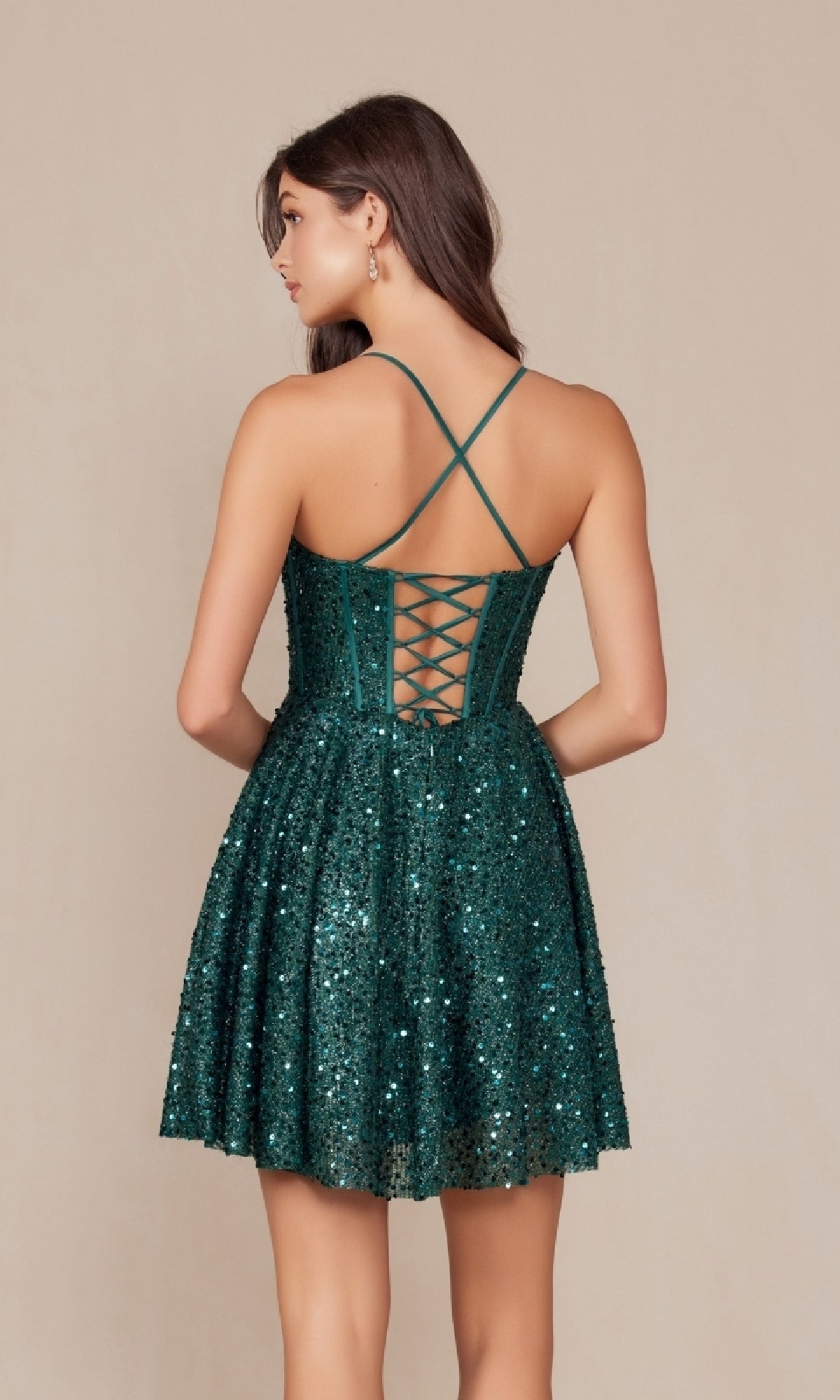 Woman in a short green Sequin Corset Homecoming Dress (Nox Anabel A843) with spaghetti straps and a lace-up back, standing against a plain beige background.